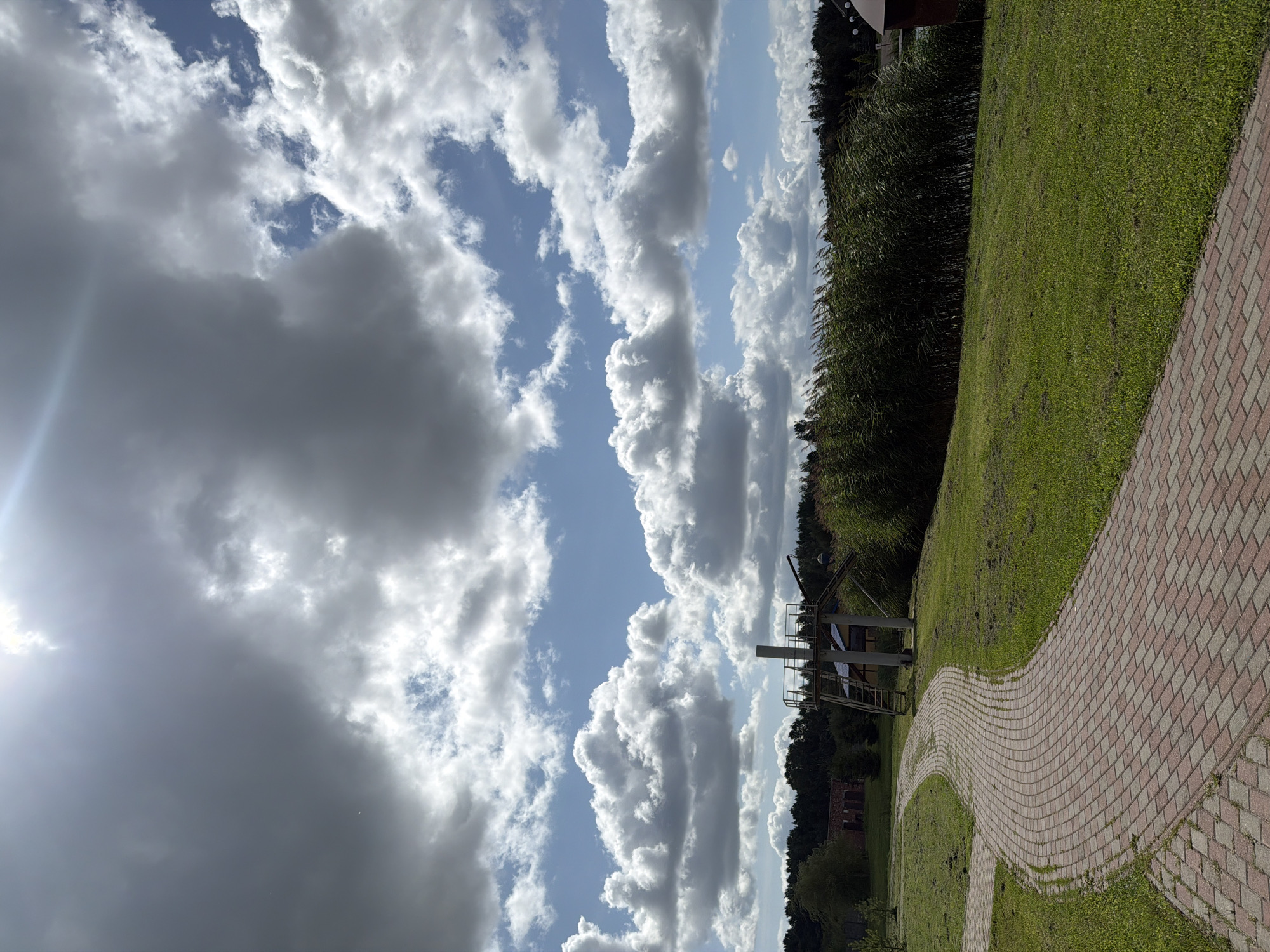 A brick walkway curves through a grassy area under a partly cloudy sky. The walkway, made of alternating red and gray bricks, leads from the lower left corner of the frame towards the middle ground. To the right of the walkway is a lawn, and further back, a dense line of tall, green reeds or marsh grass stretches across the middle and right of the image. Behind the reeds, trees form a dark green treeline. In the distance, to the left, a building with a red roof is visible, and to the right, a small portion of a red building with a white dome-like structure is seen. A tall metal structure with stairs and a platform stands on the lawn near the reeds. The sky above is filled with large, fluffy white and gray clouds, with patches of blue sky visible. The sun is bright, creating lens flare in the upper left portion of the sky.