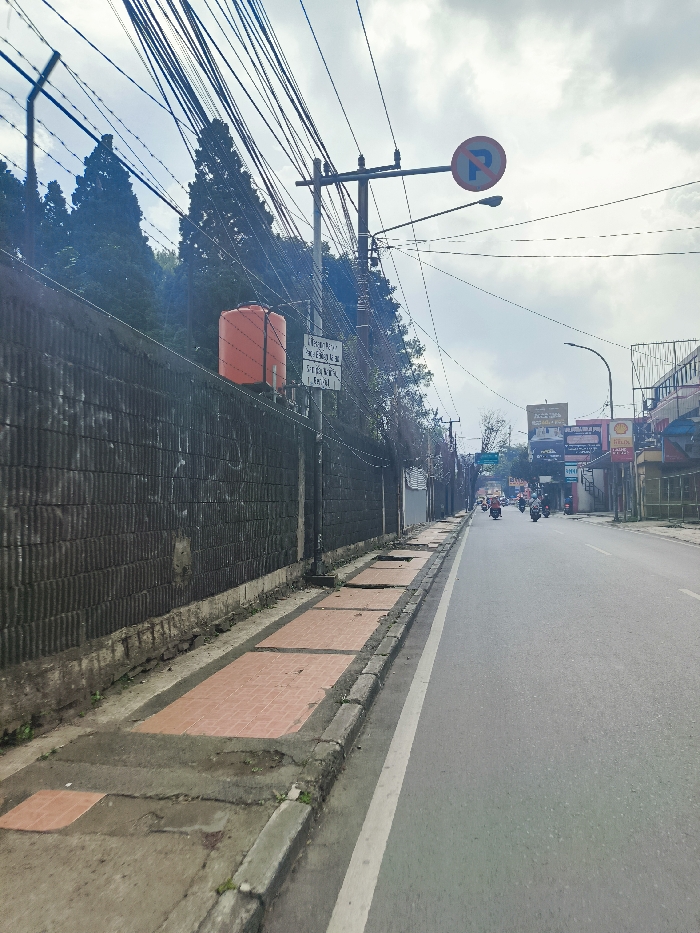 The scene is a street in Lembang, Indonesia, located at latitude -6.816 and longitude 107.616. It is a paved, two-lane road with a sidewalk on the left side, viewed from a position on the sidewalk, facing the opposite direction of traffic. The road is relatively empty with a few vehicles visible, including a motorbike, and a few figures in the distance. The sidewalk has a red tile pattern, but there are some broken sections and missing tiles. On the left side of the street, there is a high, black, vertical wall made of corrugated metal. There are multiple power lines stretching across the image, along with several utility poles. A bright orange-red water tank is mounted on a pole, located in the center of the image, beside a sign with text in Indonesian. A street sign with a white "P" in a blue circle on a white background is visible on a pole across the street. The sky is overcast, with some small clouds visible.