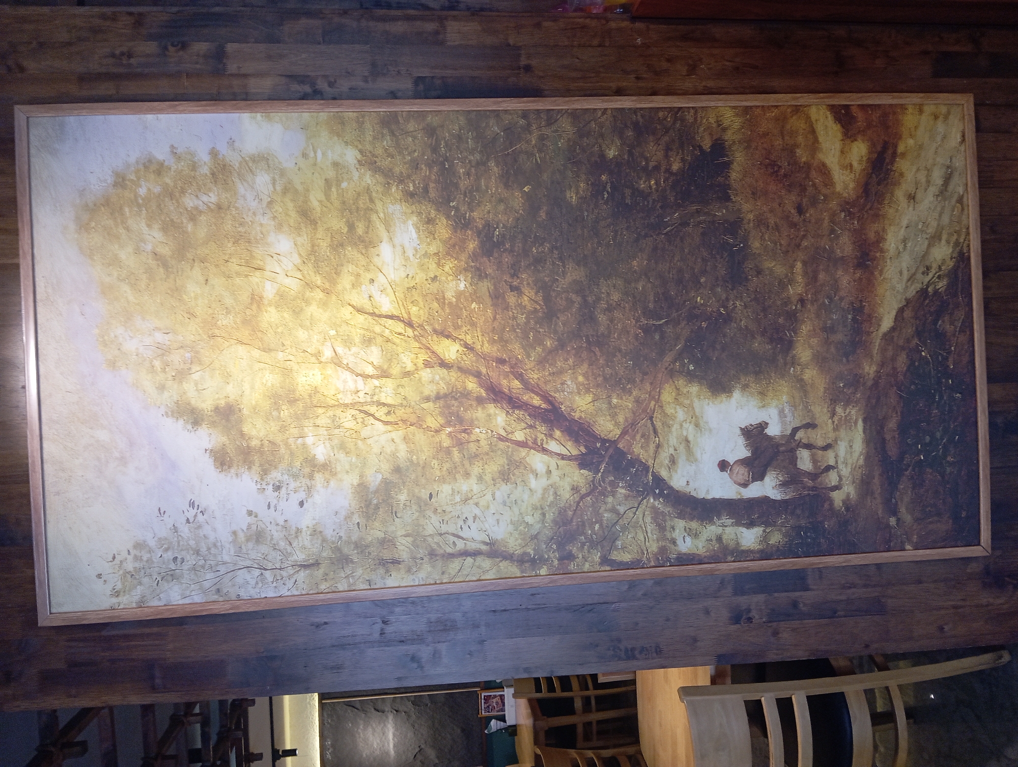 A framed painting depicting a pastoral scene hangs on a wooden paneled wall. The painting shows a light-colored horse with a rider wearing a dark jacket and hat, moving along a dirt path through a wooded area. Sunlight streams through the trees, illuminating the scene with a warm, golden glow. The overall style is impressionistic with visible brushstrokes. To the left of the painting, a chair with a light wooden frame and dark seating is visible, partially obscuring a wooden table. In the background, several other wooden chairs and tables are arranged, suggesting a dining or cafe setting. The wooden paneled wall itself appears to be made of dark, weathered planks. The location is a cafe or restaurant in Nha Trang, Vietnam.