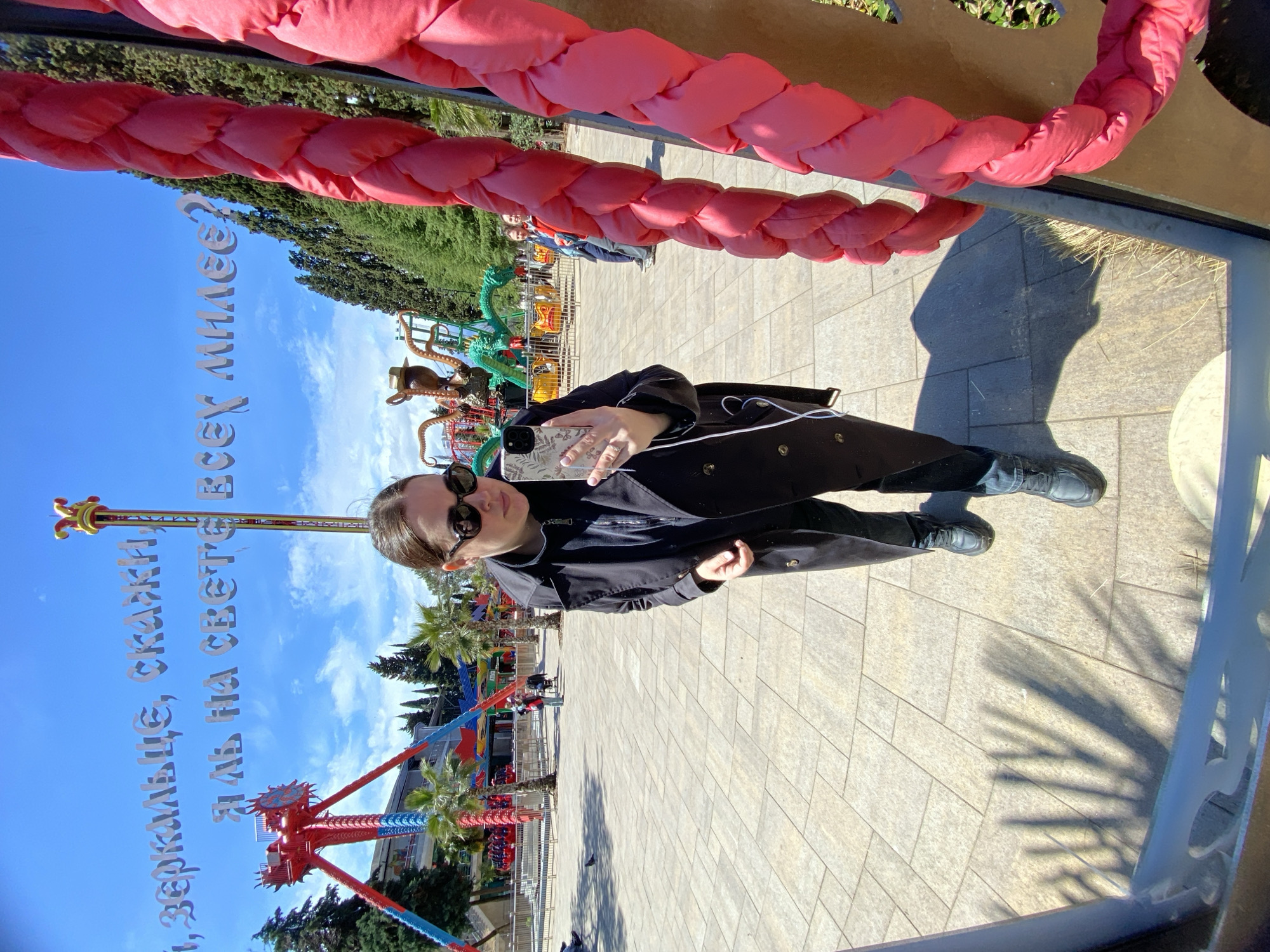 A person wearing a black coat and sunglasses stands on a paved area. In the background are amusement park rides, palm trees, and other trees. Metal lettering is visible. A padded pink rope is in the foreground. The location is likely an amusement park in Sochi, Russia.