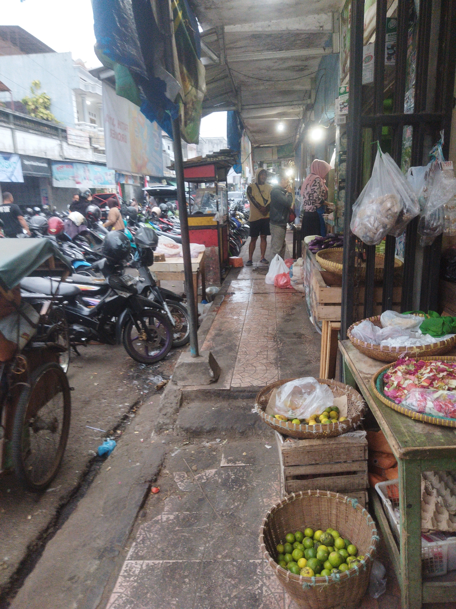 The image depicts a bustling outdoor market scene in Banjar, Indonesia, likely on a street or narrow alleyway. The market is lined with stalls covered by tarps and corrugated metal roofs, creating a covered walkway. Various goods are displayed, including baskets of fresh produce, such as limes, and what appears to be dried goods or snacks in plastic bags hanging from a stall. Several motorcycles are parked along the walkway, indicating a common mode of transportation in the area.
In the foreground, a traditional pedicab, a three-wheeled bicycle taxi, is visible on the left side of the image, its wheel prominently featured. The ground is a mixture of paved tiles and dirt, with some debris scattered around, suggesting a well-used public space.
People are present in the market, engaged in various activities. Some individuals are seen standing and interacting, possibly customers or vendors. One person in the mid-ground is wearing a pink hijab and a patterned top, appearing to be engaged in commerce. Another person, wearing a yellow jacket and dark shorts, stands with another individual dressed in a gray jacket and pants. Further in the background, more people can be seen amidst the parked motorcycles.
The overall impression is one of a lively and somewhat crowded local market, characteristic of urban life in Banjar, Indonesia. The lighting suggests it might be overcast or early morning/late afternoon, as there are no harsh shadows. The presence of plastic bags, waste, and weathered structures adds to the authentic, everyday feel of the scene.