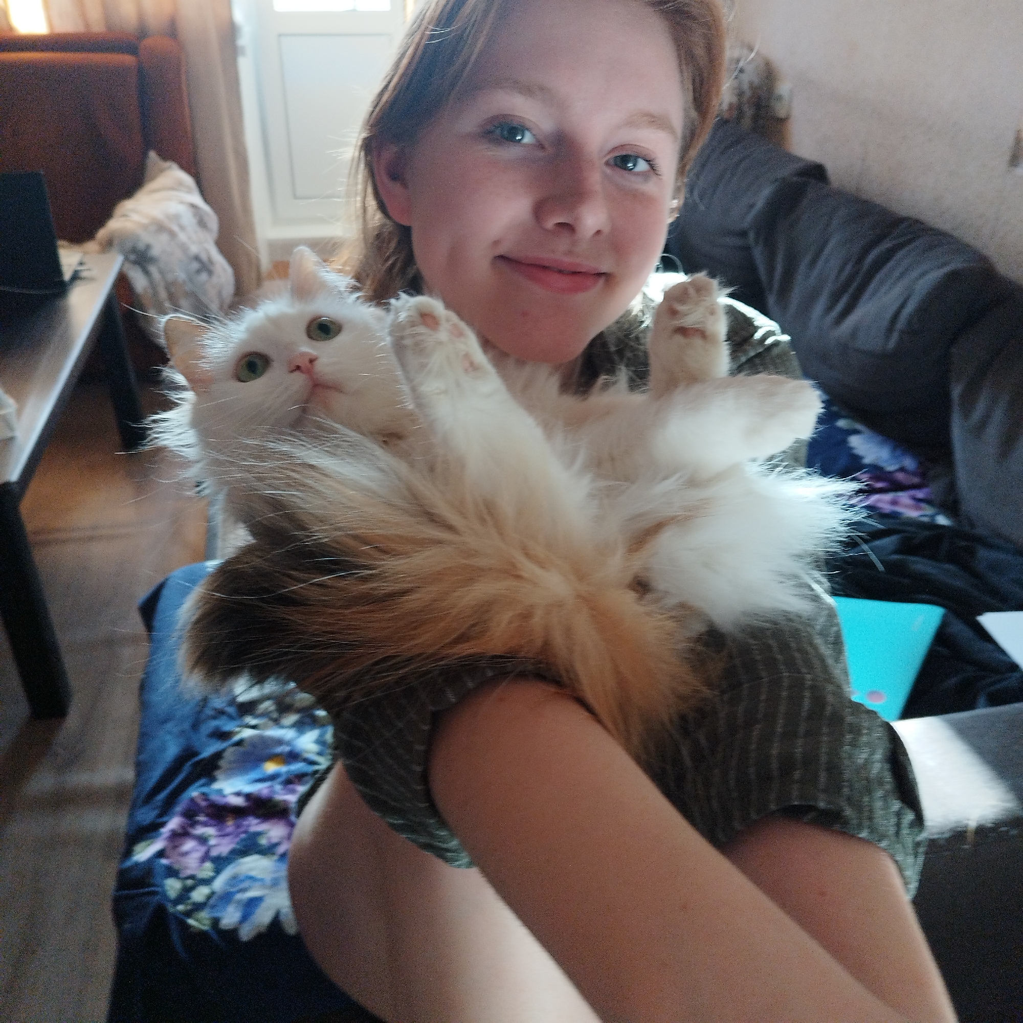 A young, light-skinned woman with reddish hair is holding a long-haired white cat with orange markings. The cat is on its back with its paws in the air. They are sitting on a floral-patterned surface with a gray couch in the background. A dark wooden table with a dark object on top is visible to the left. A window is visible in the background. The location of the photo, Yekaterinburg, Russia, is not discernible from the image.