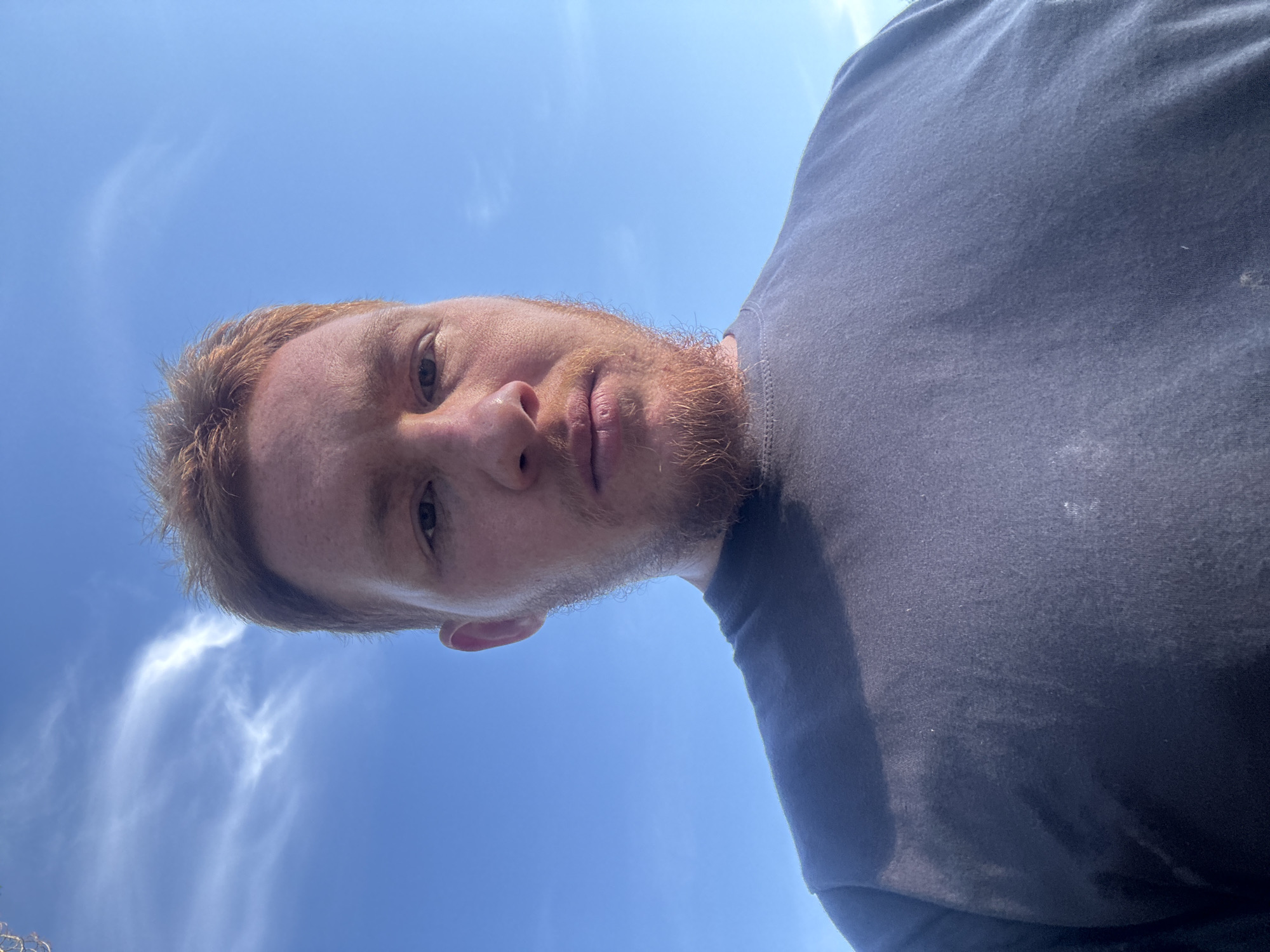 A close-up, low-angle selfie shows a white man with red hair and a beard against a bright blue sky with wispy white clouds. He is wearing a dark gray t-shirt. The man is looking directly at the camera. The background suggests an outdoor setting.