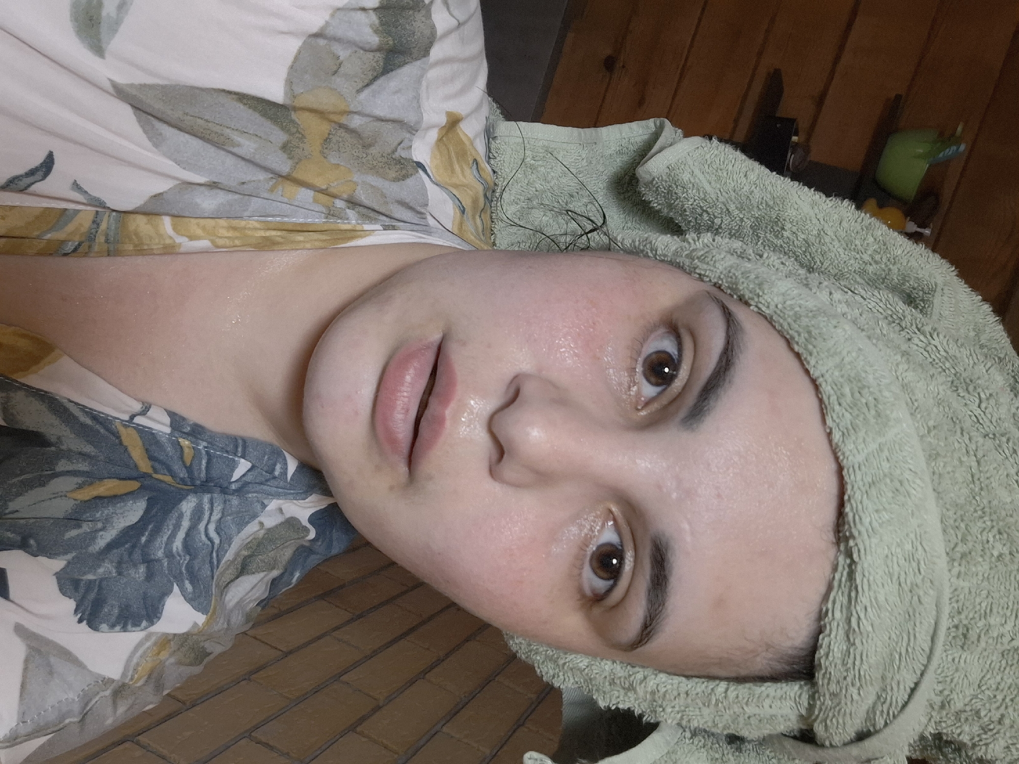 A close-up, tilted perspective captures a person with fair skin, possibly female, with a green towel wrapped around their head. The person has visible pores and a slight sheen on their face, suggesting recent cleansing or a facial treatment. Their eyes are open, and the focus is sharpest on their face. They are wearing a floral-patterned garment. The background includes a wooden surface. There is no information to indicate that this image was taken in Амзя, Russia.