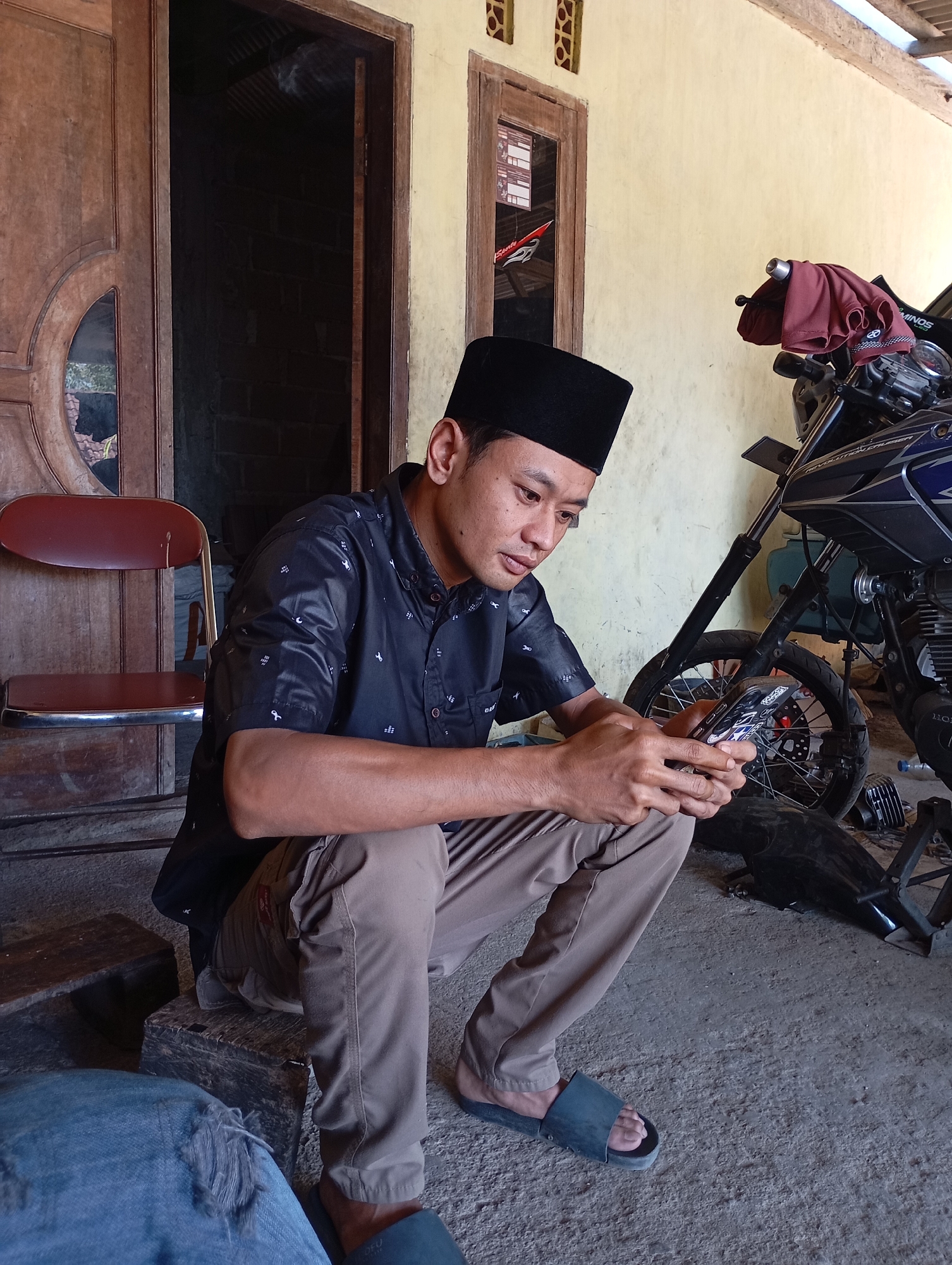 An Indonesian man wearing a black peci hat, a black patterned short-sleeved shirt, and brown pants is sitting on a wooden stool in front of a building. He is looking down at his phone, which he is holding in both hands. To his right, there is a motorcycle, and to his left, there is a reddish-brown chair. The building appears to be a house with a wooden door and a light-colored wall. The ground is concrete. The scene is outdoors, possibly in a yard or driveway, in Ketepung, Indonesia. The lighting suggests it is daytime.