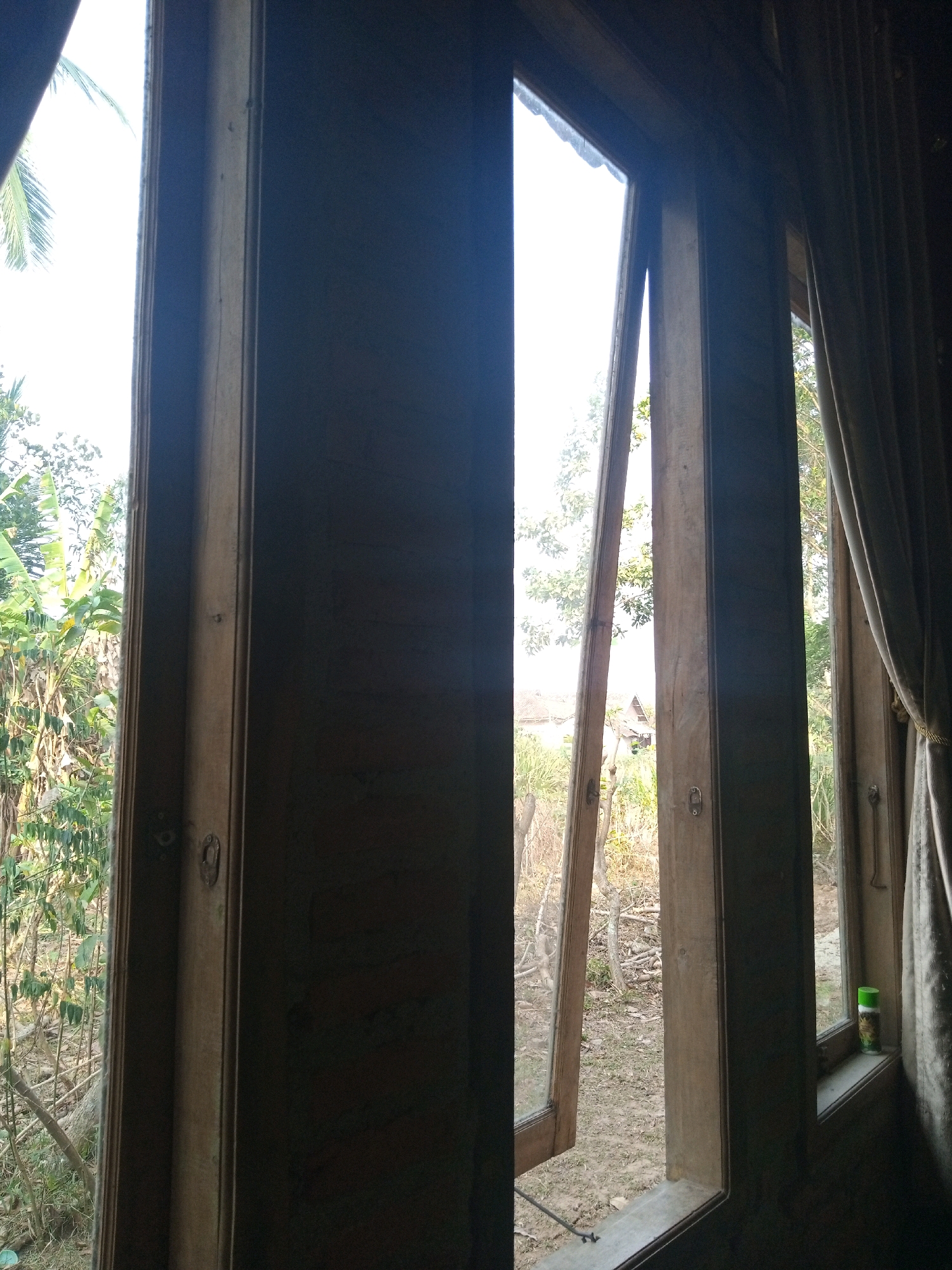 The image depicts an interior view through a wooden window frame, looking outwards at a tropical outdoor environment. The window appears to be made of dark wood and has multiple panes. One section of the window is open, allowing a clear view of the vegetation and a small building in the distance. The exterior scene features lush green foliage, trees, and a dirt ground. There are also hints of structures and what looks like a small, rustic house. The lighting suggests it is daytime. A bottle, possibly a spray can, is visible on the windowsill of another section of the window. The overall impression is one of a simple, rural dwelling in a tropical setting.