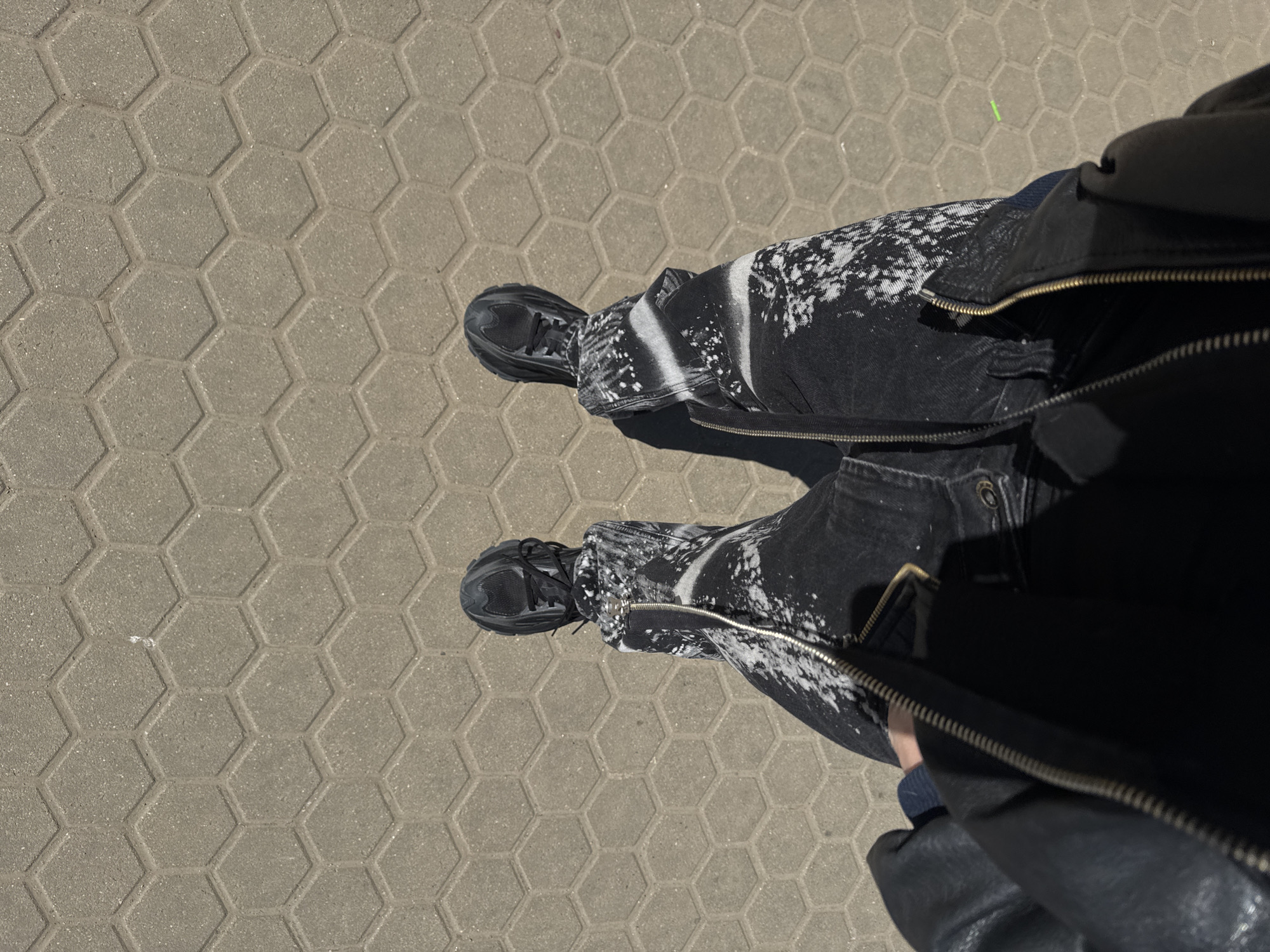 A first-person perspective photograph shows a person standing on a paved surface, presumably in an urban environment. The pavement consists of grey interlocking hexagonal blocks. The person is wearing black sneakers and black jeans with a splatter pattern on the thighs and lower legs. The jeans have zippers on the sides. A black leather jacket is partially visible in the foreground. The location appears to be outdoors in daylight, and based on the paving stones and the style of the clothing, this photograph could potentially be from Sofia, Bulgaria, though this is only speculative. There are no other visible people or objects in the frame.