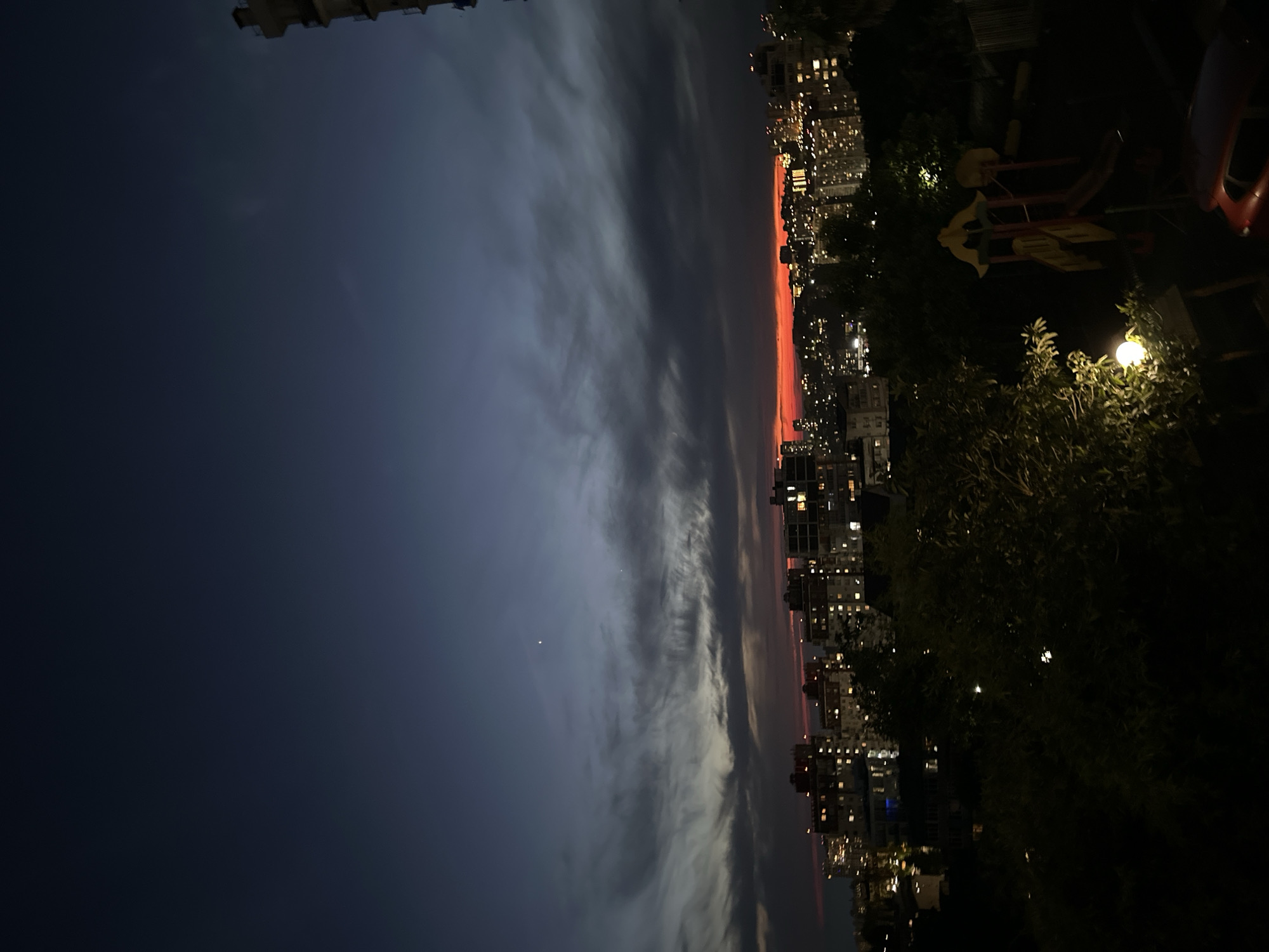 This is a photograph taken from a high vantage point, likely an apartment building, overlooking a cityscape at dusk. The sky is a deep, dusky blue, with streaks of pale clouds tinged with the last remnants of a vibrant orange-red sunset near the horizon. Several planets are visible as bright points of light in the sky.
Below the sky, a dense collection of multi-story buildings with illuminated windows stretches across the horizon. These buildings appear to be apartment complexes or office buildings, indicating a developed urban area. In the foreground, the view is partially obscured by dark silhouettes of trees and foliage. A children's playground with a red and yellow slide structure is visible to the right of the frame, and a street lamp with a glowing white orb illuminates the lower left portion of the image. A portion of a red car is also visible in the bottom right corner. The overall impression is one of a quiet, residential urban evening in Sochi, Russia.