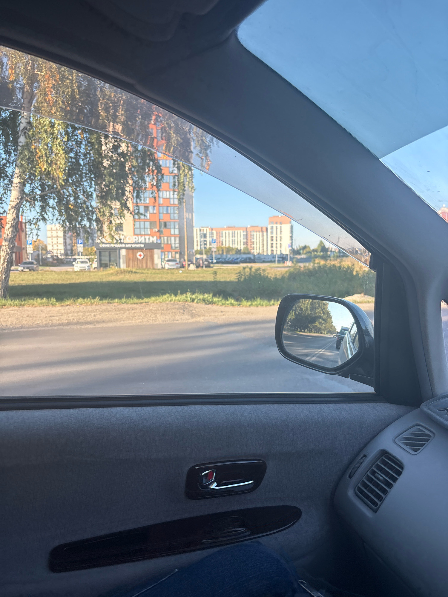 The image shows a view from inside a car, looking out the passenger side window. The scene appears to be in Barnaul, Russia, on a sunny day.
Outside the car, there is a paved road with cars driving on it, and a grassy verge. In the background, there are modern apartment buildings and some trees, one of which has autumn foliage. A small kiosk with a red cross symbol is visible on the side of the road. The side mirror of the car reflects the road ahead, showing a few other vehicles.
Inside the car, the door panel is visible, with a door handle and a storage compartment. A person's leg wearing blue jeans is visible in the lower part of the frame. An air vent is also visible on the right side of the dashboard.