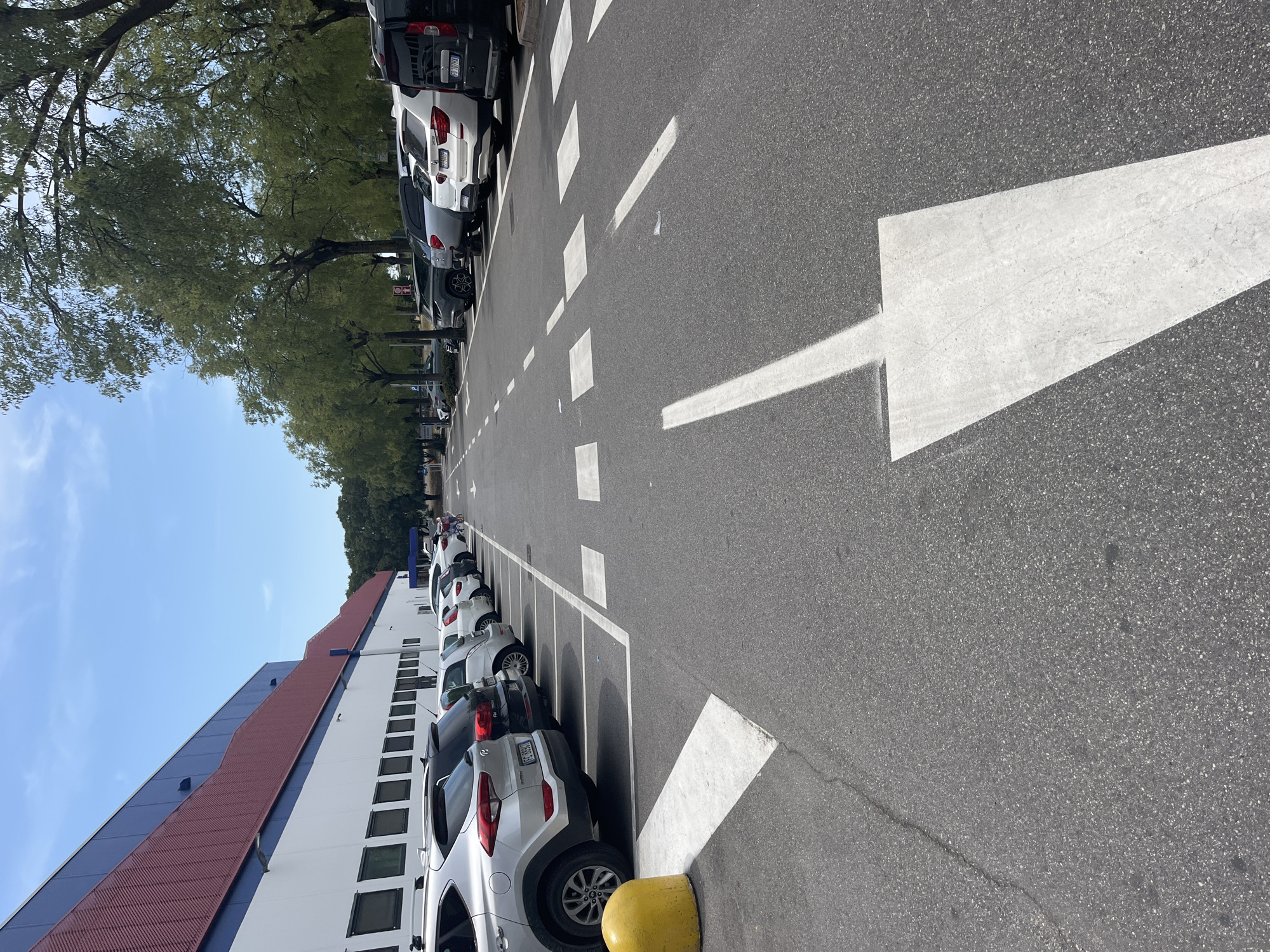 The image shows a parking lot in front of a large building with a red and blue facade. The building appears to be a commercial establishment, possibly a supermarket or warehouse. The parking lot is asphalt, with white lines marking parking spaces and directional arrows. Several cars are parked in the spaces, and some trees line the edge of the parking lot. In the distance, two people are pushing a shopping cart. The sky is blue with some clouds. This scene is consistent with the parking area of a large retail store, such as a Costco, located in Castiglione delle Stiviere, Italy.