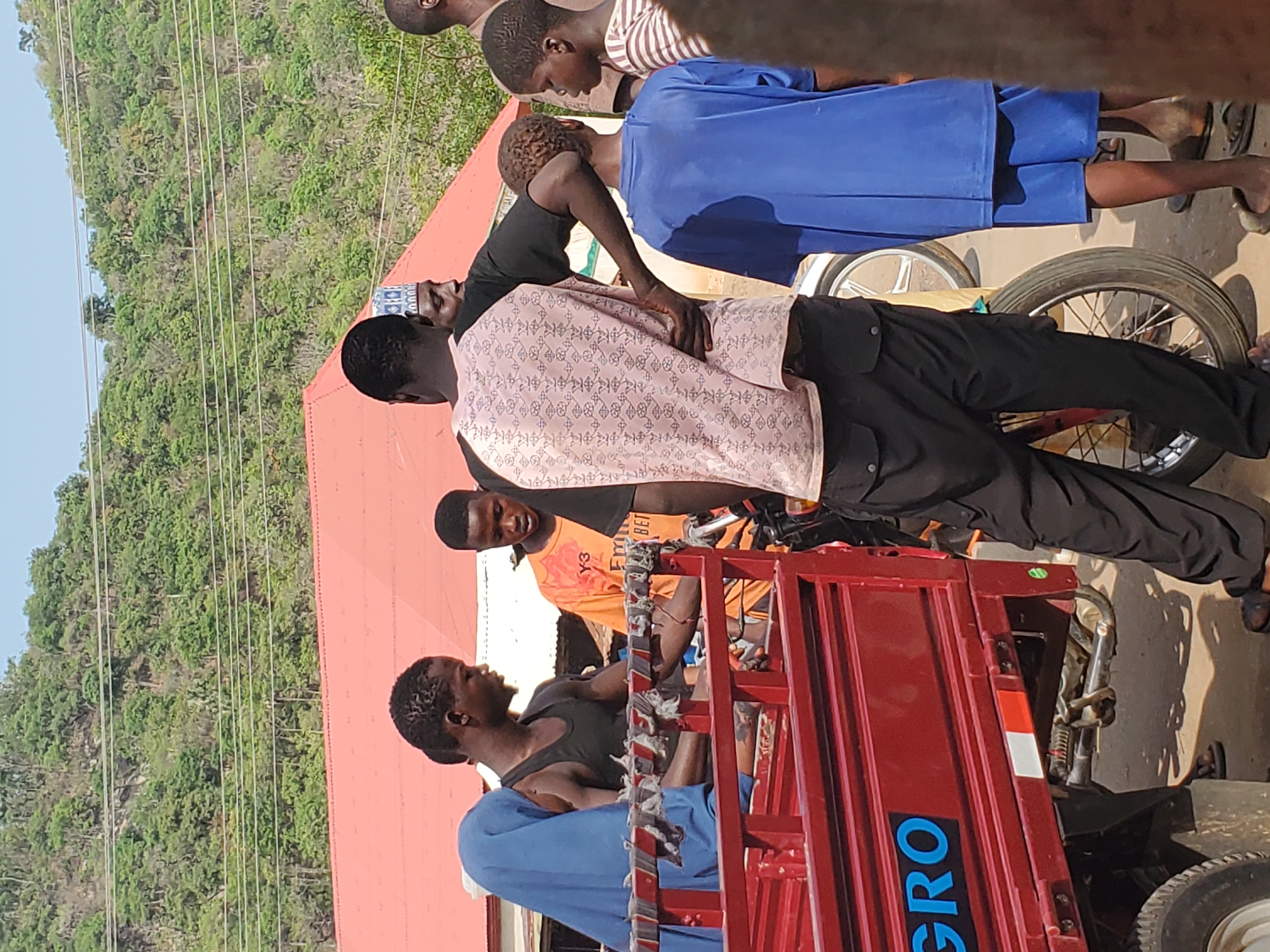 In Mararaba, Nigeria, a group of people stand around a red three-wheeled motor vehicle. A man sits in the back of the vehicle, wearing a blue garment and a black tank top. Several other individuals surround the vehicle. One man wears a light pink patterned shirt and black pants. Another is wearing an orange t-shirt. Two young people dressed in blue school uniforms are also present. The background shows a building with a red roof, green vegetation, and power lines.