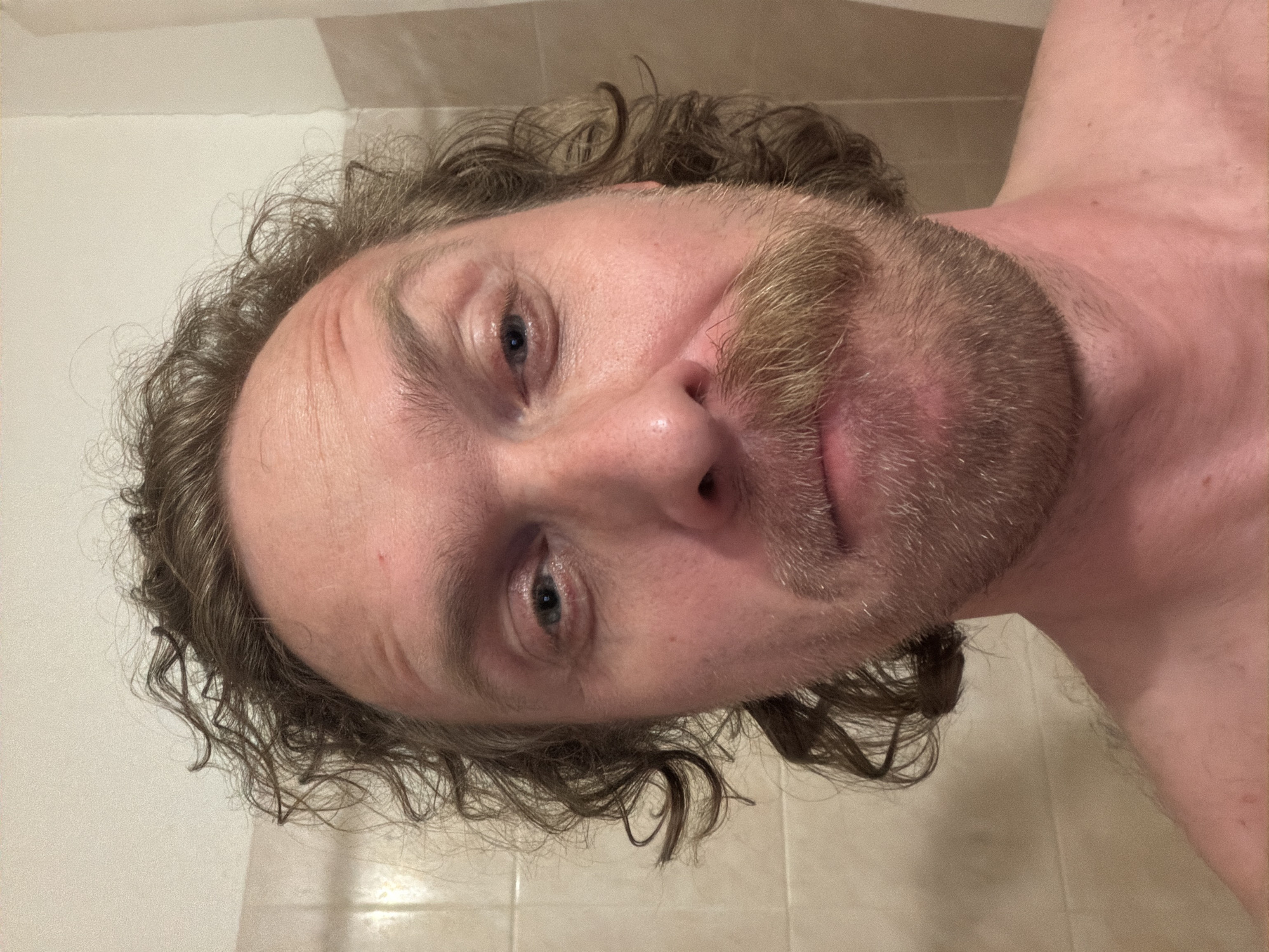 A middle-aged man with curly brown hair and a beard poses in a bathroom. The bathroom has light-colored walls and tiled floors. The location cannot be determined from the image.