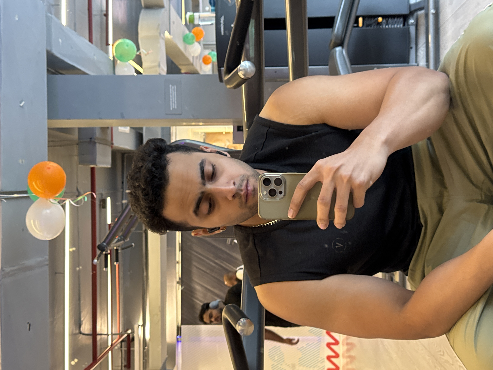 A man is taking a selfie in a gym. He is wearing a black sleeveless shirt and olive green pants. He has on a gold chain and black earbuds. In the background, there are exercise machines and other people working out. There are also balloons hanging from the ceiling, some of which are white, orange, and green. The gym appears to be modern with a high ceiling and fluorescent lighting. The overall impression is that of a well-equipped fitness facility. The user specified the city of Kolkata, India, however, there is no specific visual information in the image that can confirm this location.