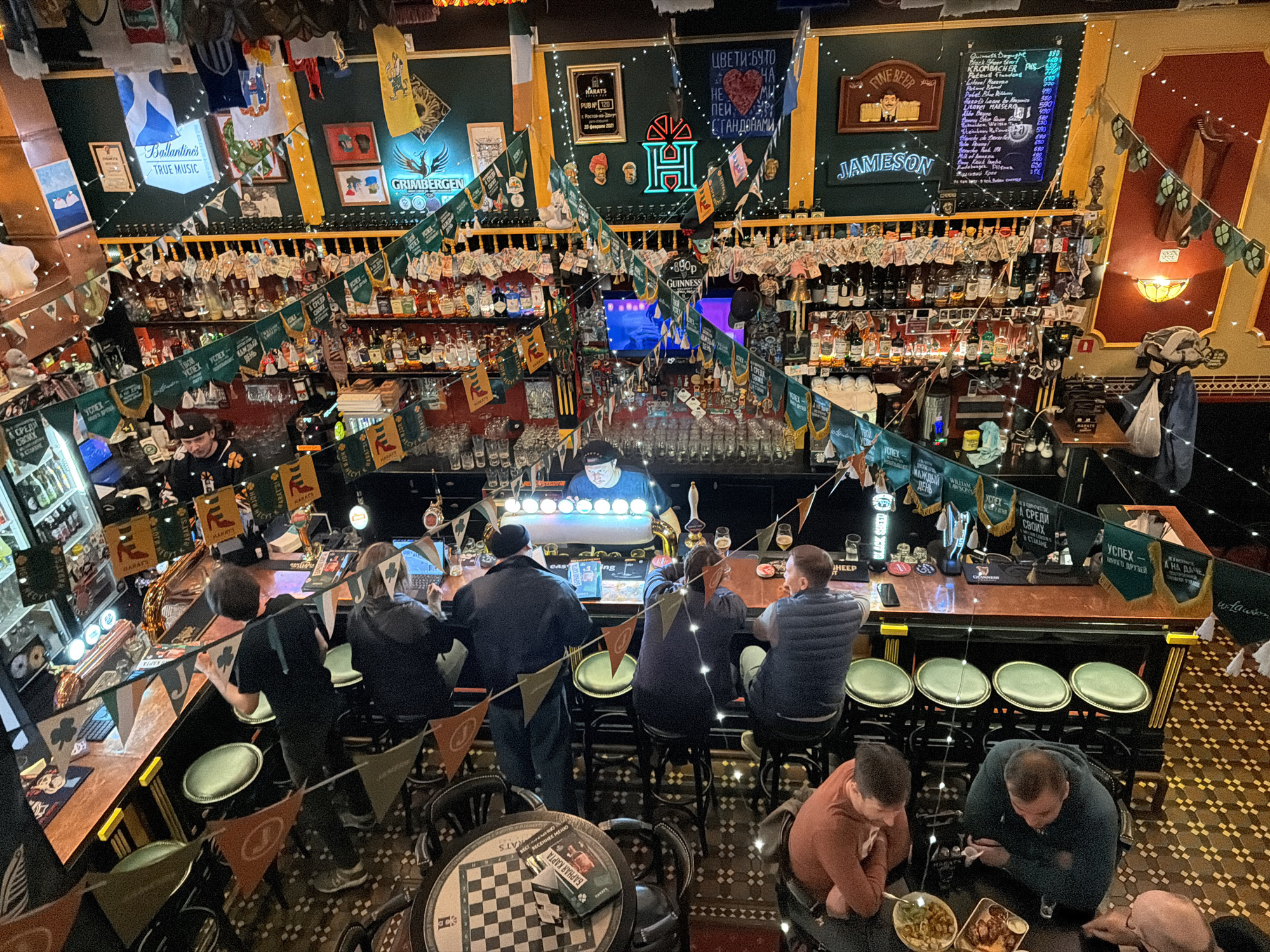 A high-angle shot of the interior of Harat's Pub in Rostov-on-Don, Russia. Customers are seated at the bar and tables, consuming food and drinks. The bar is well-stocked, with a wide range of beverages displayed. The pub is decorated with Irish-themed decor and banners. A sign indicates a menu with pricing. The floor features an ornate tile pattern.