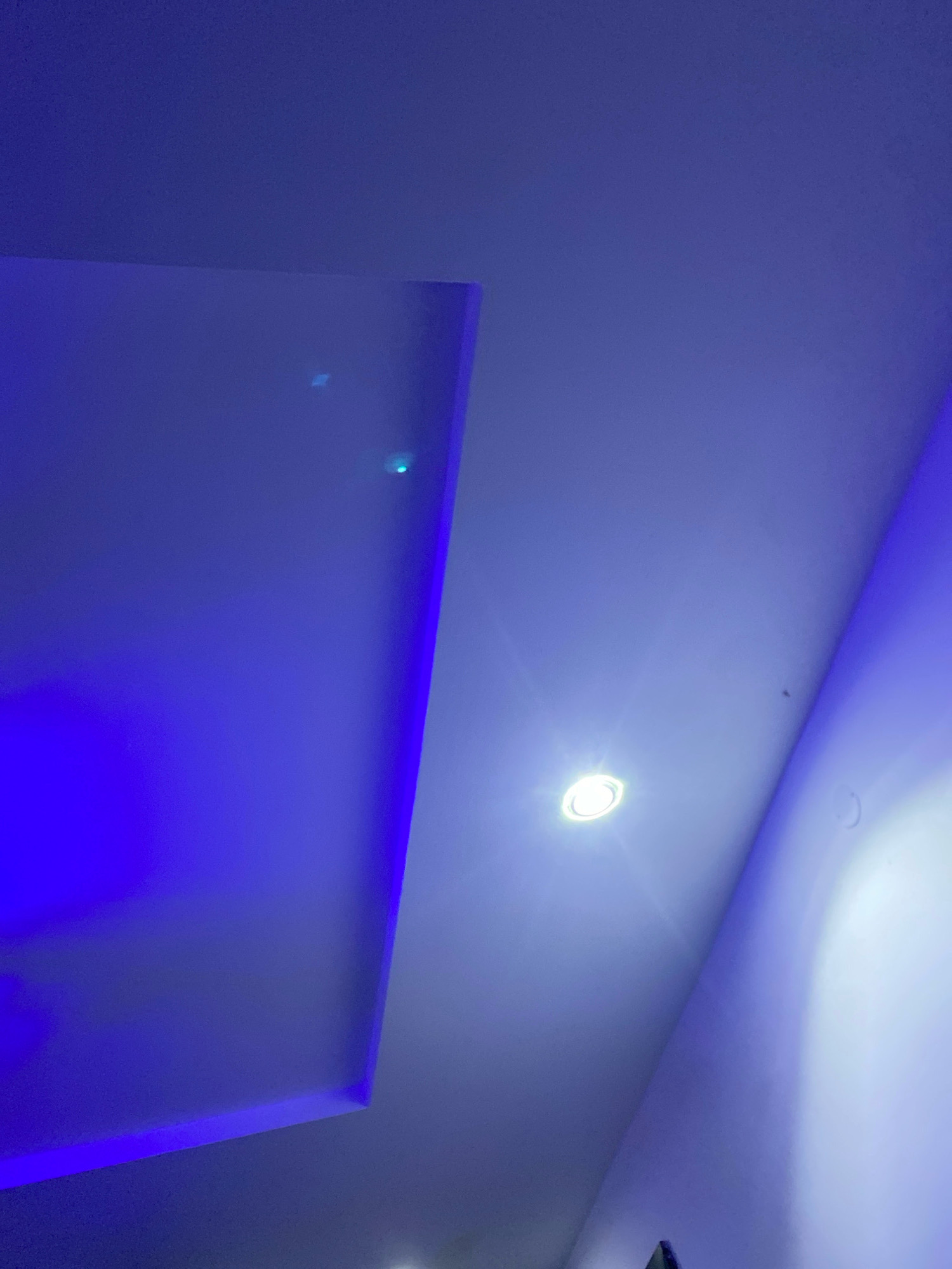 The image shows a ceiling with blue ambient lighting and a single white recessed light fixture. The ceiling appears to be made of plasterboard and has a stepped design in the corner. There is a rectangular section of the ceiling illuminated by a bright blue light, while another section is lit by the white spotlight. The environment appears to be indoors, possibly a room in a residential or commercial building. There are no people or specific objects visible that would indicate a location in Shangisha, Nigeria.