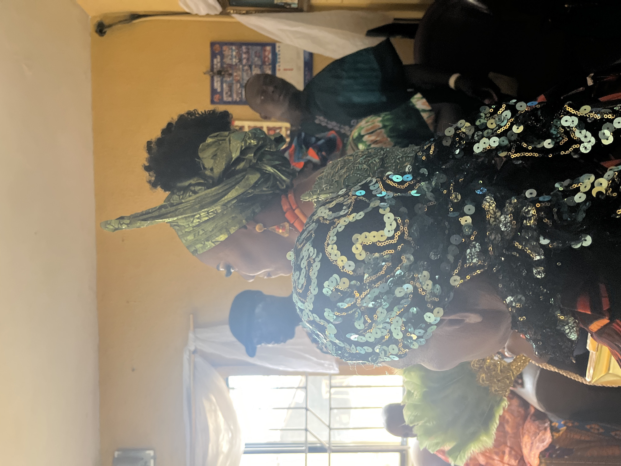 In an indoor setting in Warri, Nigeria, three individuals are present. The woman on the left wears a green head wrap, while the woman on the right has a black head covering decorated with pale green sequins. The man in the background is partially visible, wearing a dark green shirt with a colorful patterned design. A window is visible in the background, letting in bright sunlight. The wall is painted a muted yellow. A framed object hangs on the wall behind the man.