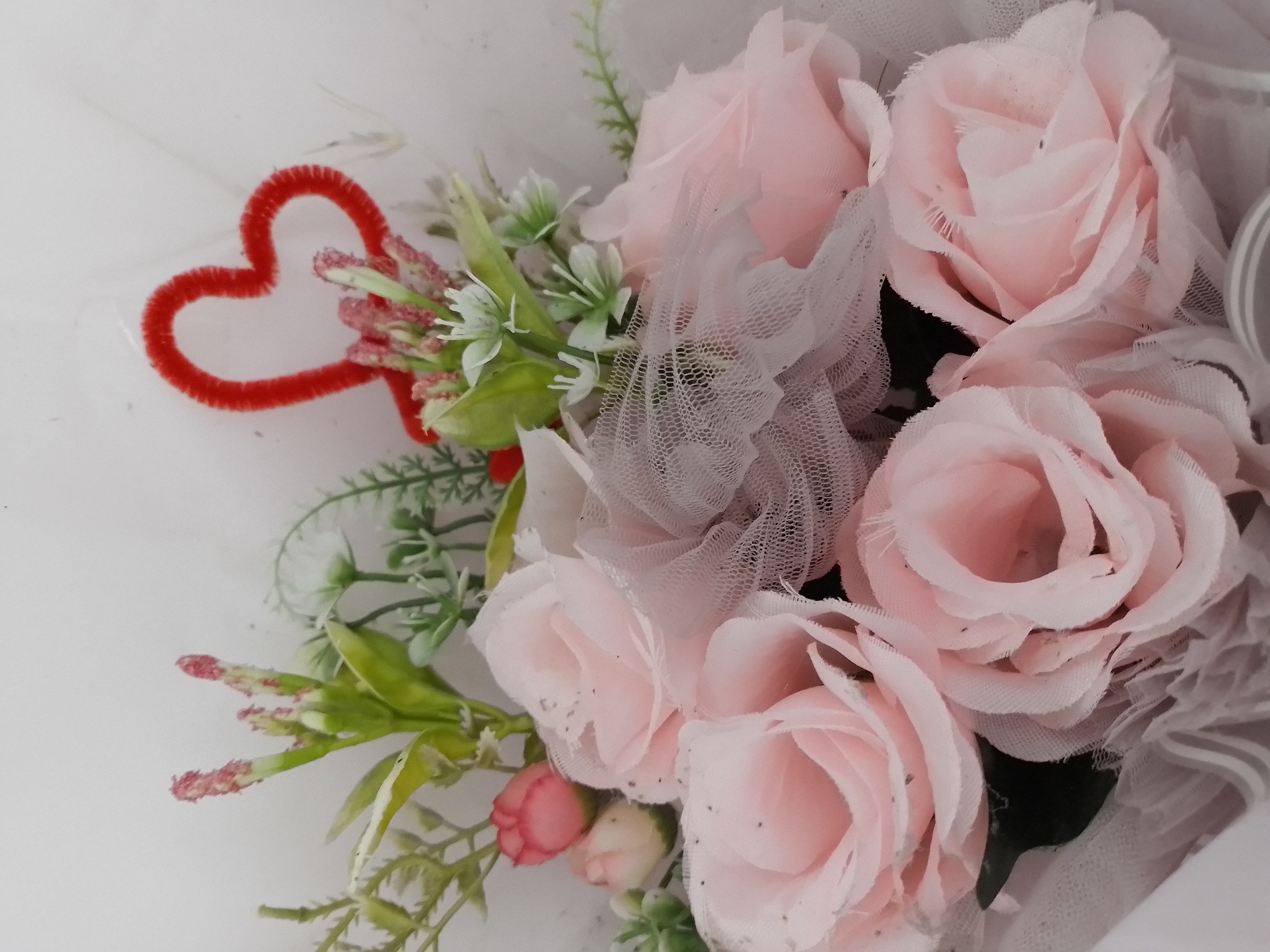 A close-up, overhead shot shows a bouquet of light pink artificial roses and greenery, adorned with a red pipe cleaner heart and white tulle. The flowers are layered and arranged in a circular pattern, with the tulle draped around them. The background is a plain white surface. The image is sharp and well-lit, focusing on the texture and detail of the bouquet. The roses appear to be made of fabric, with delicate, layered petals. The greenery is a mix of artificial leaves and sprigs of small white flowers. A thin red pipe cleaner, shaped like a heart, is visible at the top of the bouquet. White tulle is wrapped around the flowers, adding a soft, airy quality to the arrangement. The overall impression is one of a charming and romantic gift.
