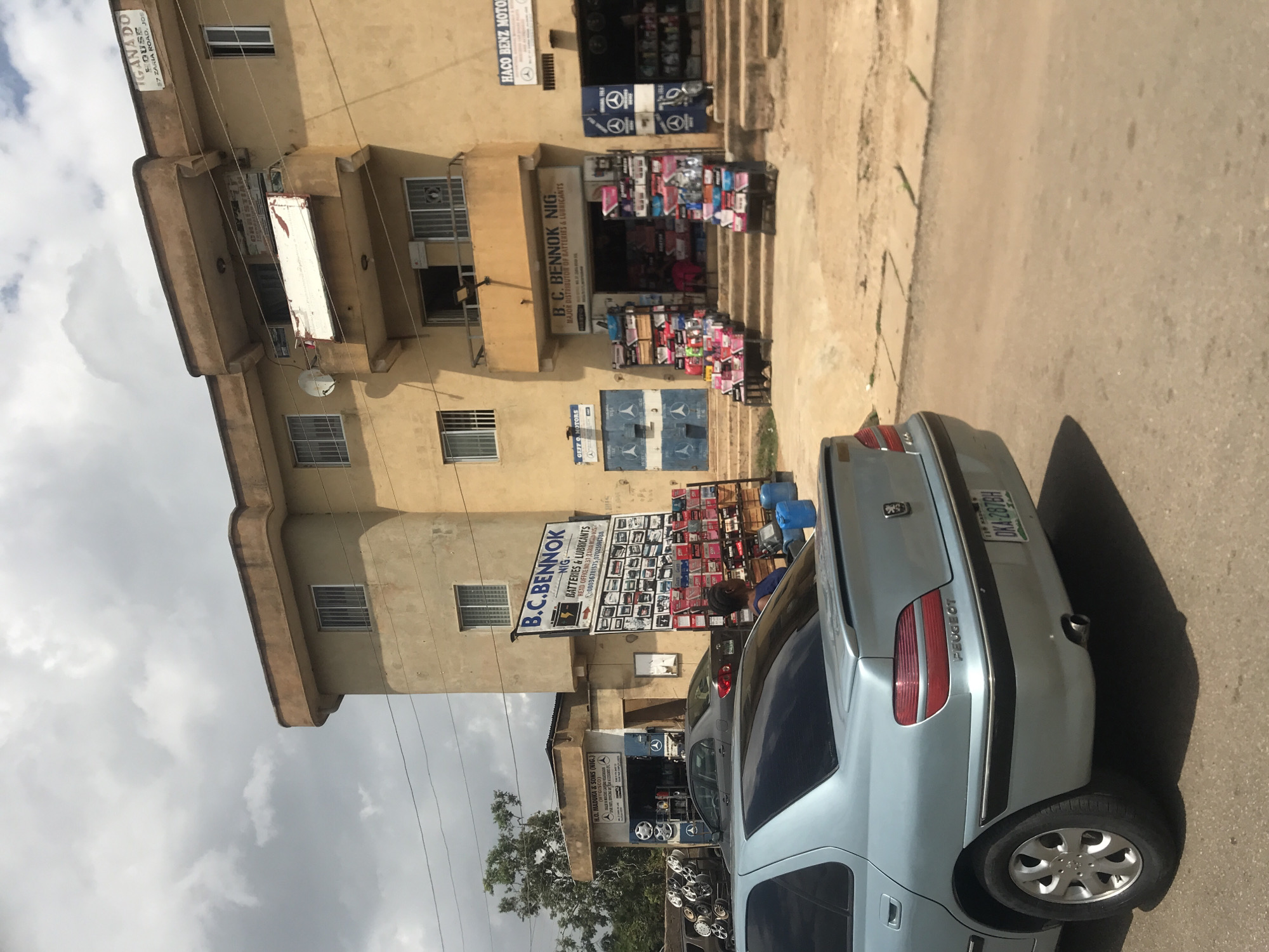 In New Stadium, Nigeria, a light blue Peugeot car is parked on a street in front of a multi-story building. The building's facade is tan, with several windows and balconies. A sign on the building reads "B.C. BENNOK NIG. MAJOR DISTRIBUTOR OF BATTERIES & LUBRICANTS". In front of the building, several displays of batteries and other automotive parts are arranged on shelves. To the left of the main building, a smaller shop with a sign that says "H.O. MADUKA & SONS (NIG.)" is visible, also displaying car parts. The street appears to be paved, with some dirt and debris present. The sky is cloudy. A woman in a blue top is standing near the batteries on display, facing away from the camera.