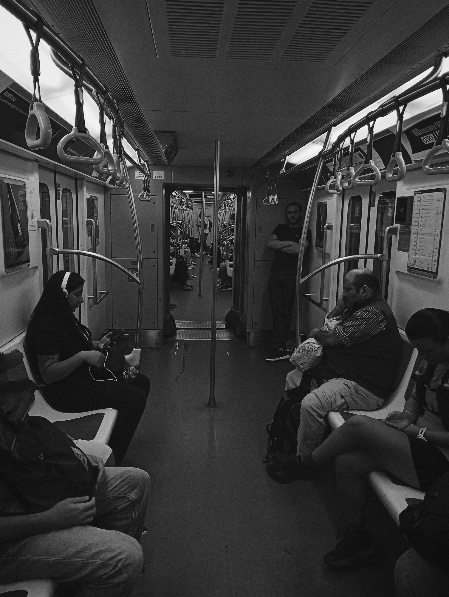The black and white image depicts the interior of a subway car in Buenos Aires, Argentina, likely from the Subte system. The environment features modern design elements: overhead fluorescent lighting, rows of white plastic bench seating, numerous light-colored grab handles suspended from horizontal bars, and dark, reflective flooring. Metal support poles are positioned throughout the carriage. Spanish signage is visible, including "MARTILLO En caso de emergencia, rompa el vidrio" (Hammer. In case of emergency, break the glass) on the left wall and "Próximo" (Next) on the right.
Approximately six individuals are clearly visible. In the foreground on the left, a female with long dark hair, wearing over-ear headphones and a dark top with visible arm tattoos, is seated. She is looking down at a smartphone connected via a cable, with a dark backpack at her feet. In the mid-ground on the right, a bald, bearded male in a plaid shirt and light pants is seated, holding an infant wrapped in a patterned blanket and looking down at the child. Adjacent to him, a female with dark hair in a dark top is seated with crossed legs, looking downwards; a smartwatch is visible on her left wrist. Standing mid-carriage, between the two groups of seated passengers, is a male with short dark hair and a beard, wearing a dark t-shirt and dark pants. He stands with his arms crossed, leaning against a door frame, facing forward. Several other passengers are partially visible further down the carriage, both seated and standing. No direct interactions between the primary subjects are apparent.