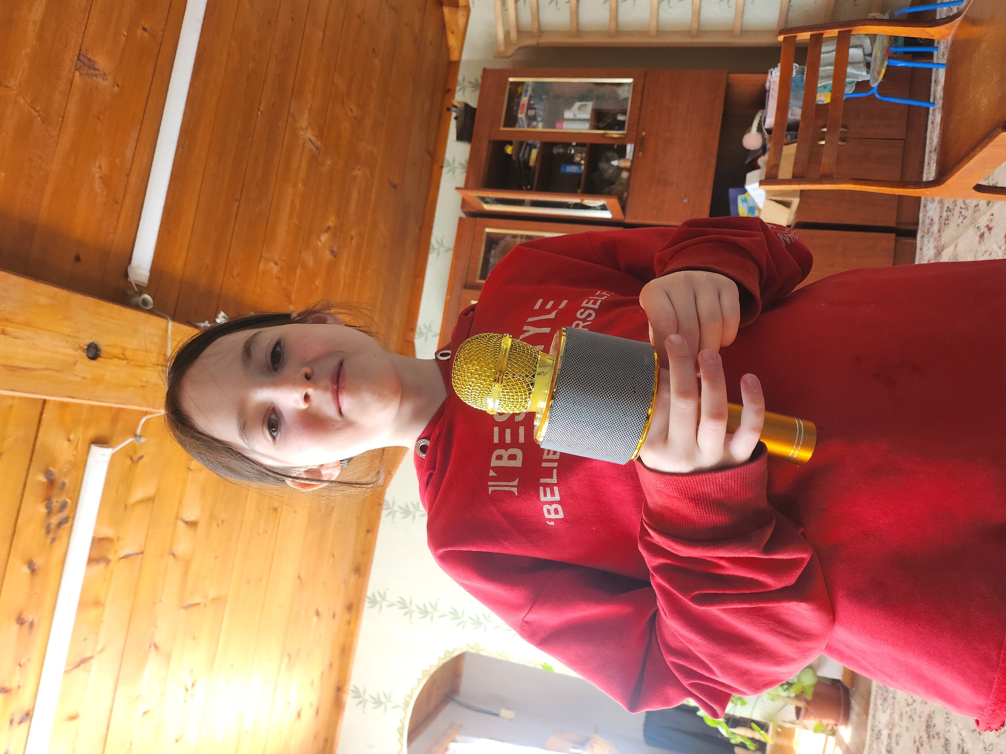 A young girl stands in a room with wood-paneled walls and ceiling, holding a gold and gray microphone. She is wearing a red hooded sweatshirt with white text that reads "I BELIEVE IN YOURSELF". Behind her is a wooden cabinet with glass doors. The room contains a wooden chair with blue accents. The location is Архангельское, Russia.