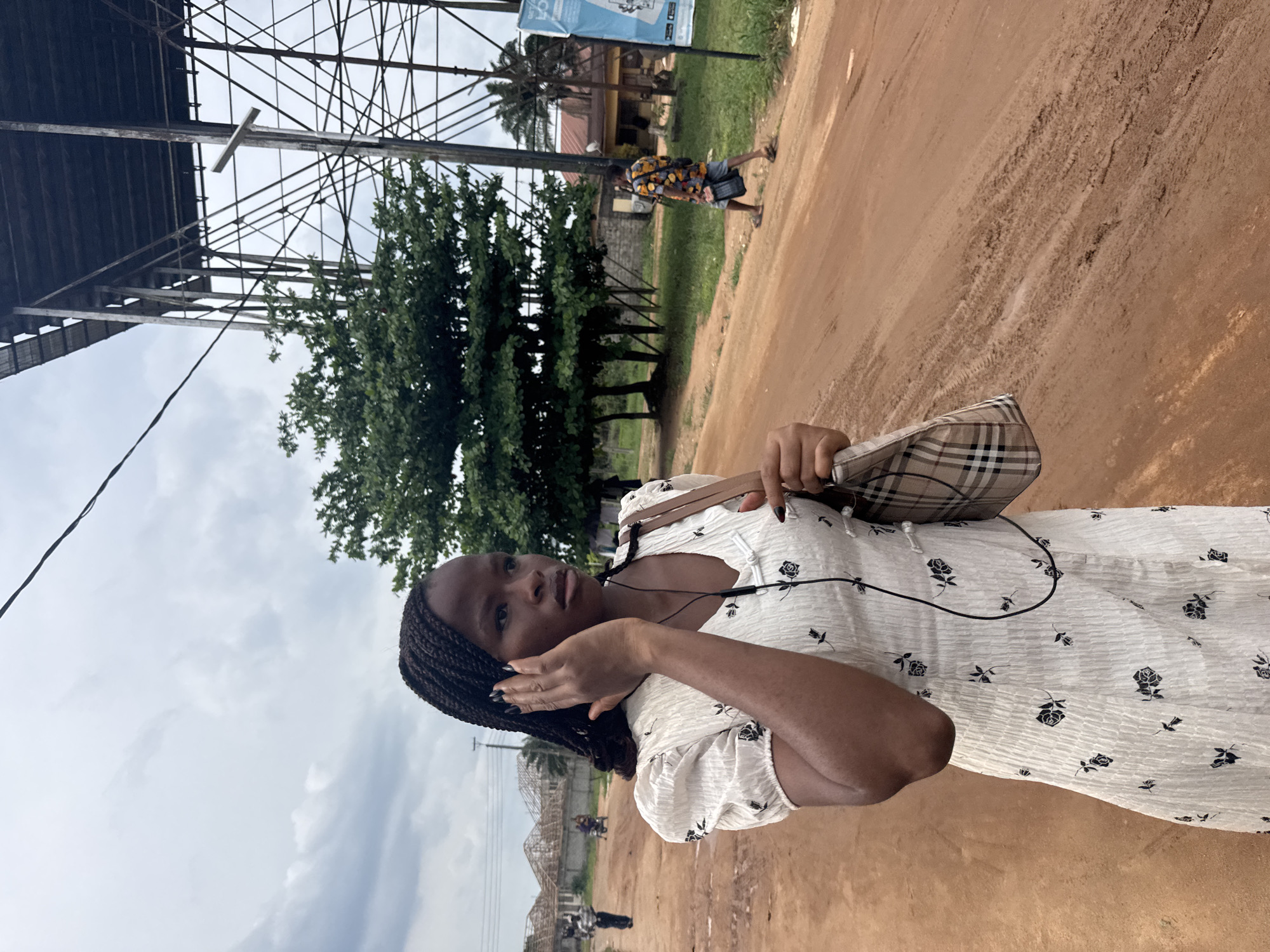 The image features a young Black woman as the main subject, positioned slightly to the right of the frame. She has braided hair and is wearing a white short-sleeved dress with dark floral patterns. She holds a plaid-patterned shoulder bag (beige, black, red, white check) and has wired earphones in her ears, with one hand raised near her ear. Her expression is neutral as she looks towards the camera.
The scene is outdoors on a reddish-brown dirt road or path, characteristic of laterite soil often found in regions like Nigeria. The ground shows some unevenness and texture. In the background, to the left, a large, complex metal framework structure, possibly part of a roof or scaffold, dominates the upper left portion of the sky. Green trees and bushes are scattered throughout the mid-ground. Another person, wearing a colorful patterned shirt and shorts, is seen walking in the mid-distance along the path. Further in the background, some low-rise buildings and power lines are visible.
The sky is overcast with greyish-white clouds, suggesting daytime. The overall setting appears to be a semi-urban or rural area in Ihiagwa, Nigeria. There is a light blue sign in the distant background with some unreadable text, though partial letters "CA" or "BLUE" might be discernible. The woman's fingernails are painted a dark color.