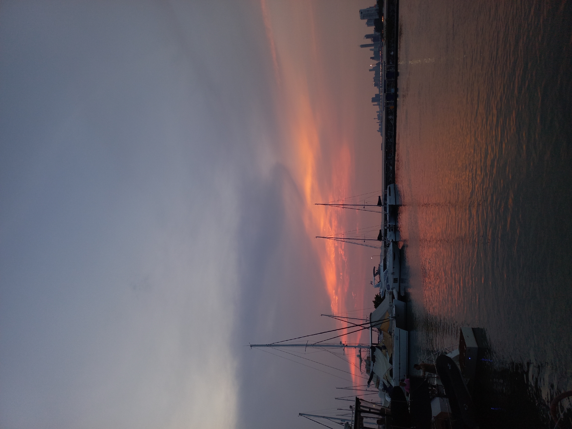 The image captures a sunset over a marina in Na Chom Thian, Thailand. The sky exhibits a gradient of colors, transitioning from a dark grey on the left to vibrant orange and pink hues near the horizon on the right. The skyline of Na Chom Thian is visible in the far right distance, with buildings silhouetted against the fading light.
Several sailboats are docked at the marina, their masts reaching towards the sky. The water reflects the colors of the sunset, creating a rippled and shimmering effect. Several small boats and inflatable dinghies are docked closer to the viewer.