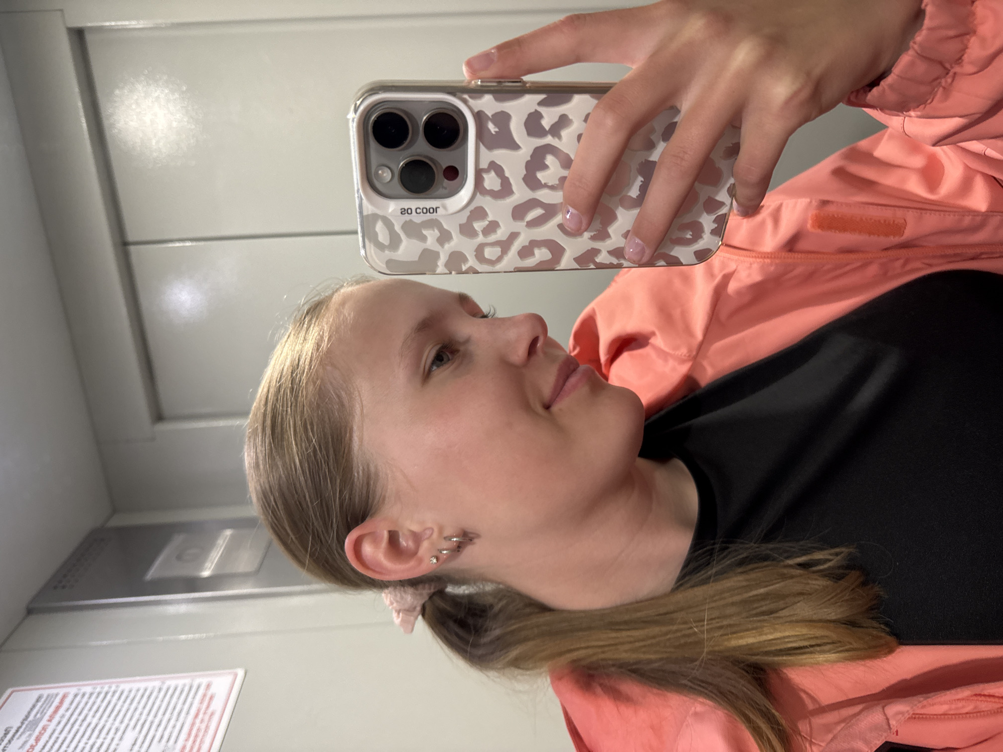 The image is a mirror selfie featuring a young woman in an indoor setting. Her face is visible in profile, looking slightly upwards with a gentle smile. She has light brown hair tied back with a pink scrunchie, and her left ear displays multiple piercings, including small hoops and a stud.
She is wearing a coral or peach-colored jacket with a quilted or puffer texture over a black top. A small orange velcro patch is visible on the inner flap of the jacket. Her right hand, with light pink polished nails, holds an iPhone in a clear case featuring a distinctive light grey leopard print pattern. The triple-lens camera module of the phone is prominent. The text "GO COOR" is visible on the phone case, appearing reversed in the mirror reflection.
The background consists of plain, light-colored walls, possibly paneled, suggesting an interior space like a hallway or elevator. At the bottom left, a blurry white sign or paper with unreadable text can be seen. The overall lighting is bright and even, consistent with an indoor environment. The location context provided for the image is Chaikovsky, Russia.