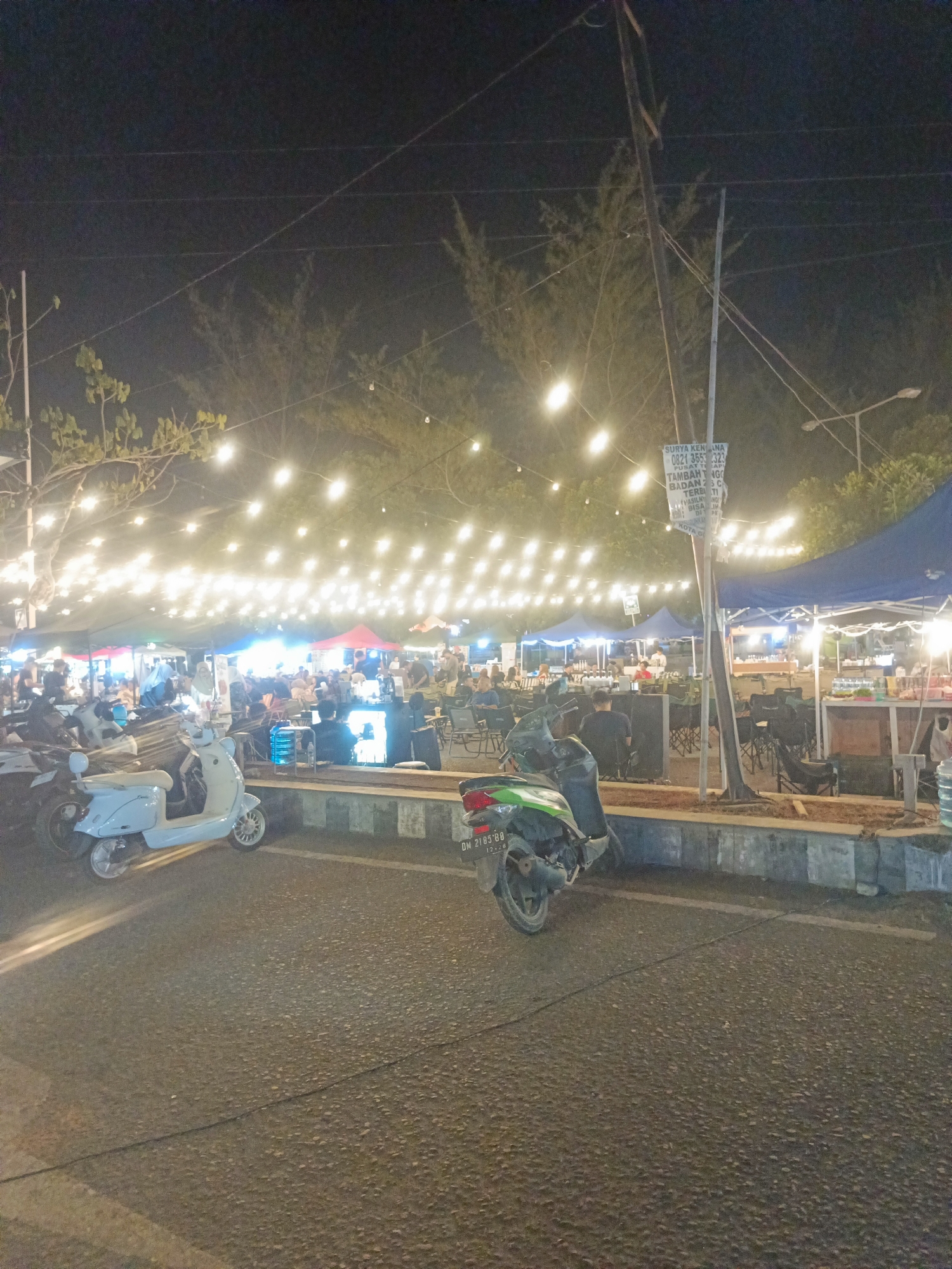 This is a night-time photograph taken at the Luwoo beach street food area in Luwoo, Indonesia. The scene depicts a bustling outdoor dining environment with numerous stalls illuminated by strings of fairy lights suspended above. Several people are visible, seated at tables and congregating around food stalls. In the foreground, two scooters are parked on the asphalt road. One scooter is light blue, and the other is dark green. The background features tents and trees. A sign with text is mounted on a pole in the middle ground. The overall atmosphere is lively and social.