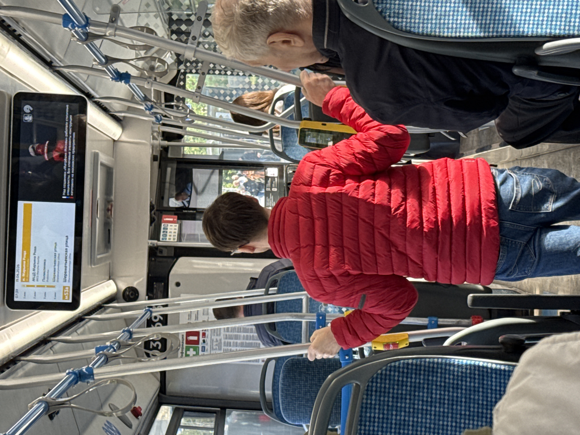 The image shows the interior of a public bus in Moscow, Russia. The camera is positioned from the perspective of a passenger seated towards the back of the bus, looking forward. In the foreground, a person wearing a red puffer jacket and blue jeans is standing and holding onto a metal pole. To their right, an older man with gray hair, also a passenger, is seated and looking forward. The bus is equipped with blue and white patterned seats and metal handrails. Above the passengers, a digital display screen shows bus route information, including the date "19.06.2020" and station names in Russian, indicating it's a Moscow bus. Another smaller screen above that shows a news report or advertisement. Sunlight streams in through the windows, illuminating the interior. Several other passengers are visible in the background, seated and obscured by the central aisle. The environment appears to be daytime.