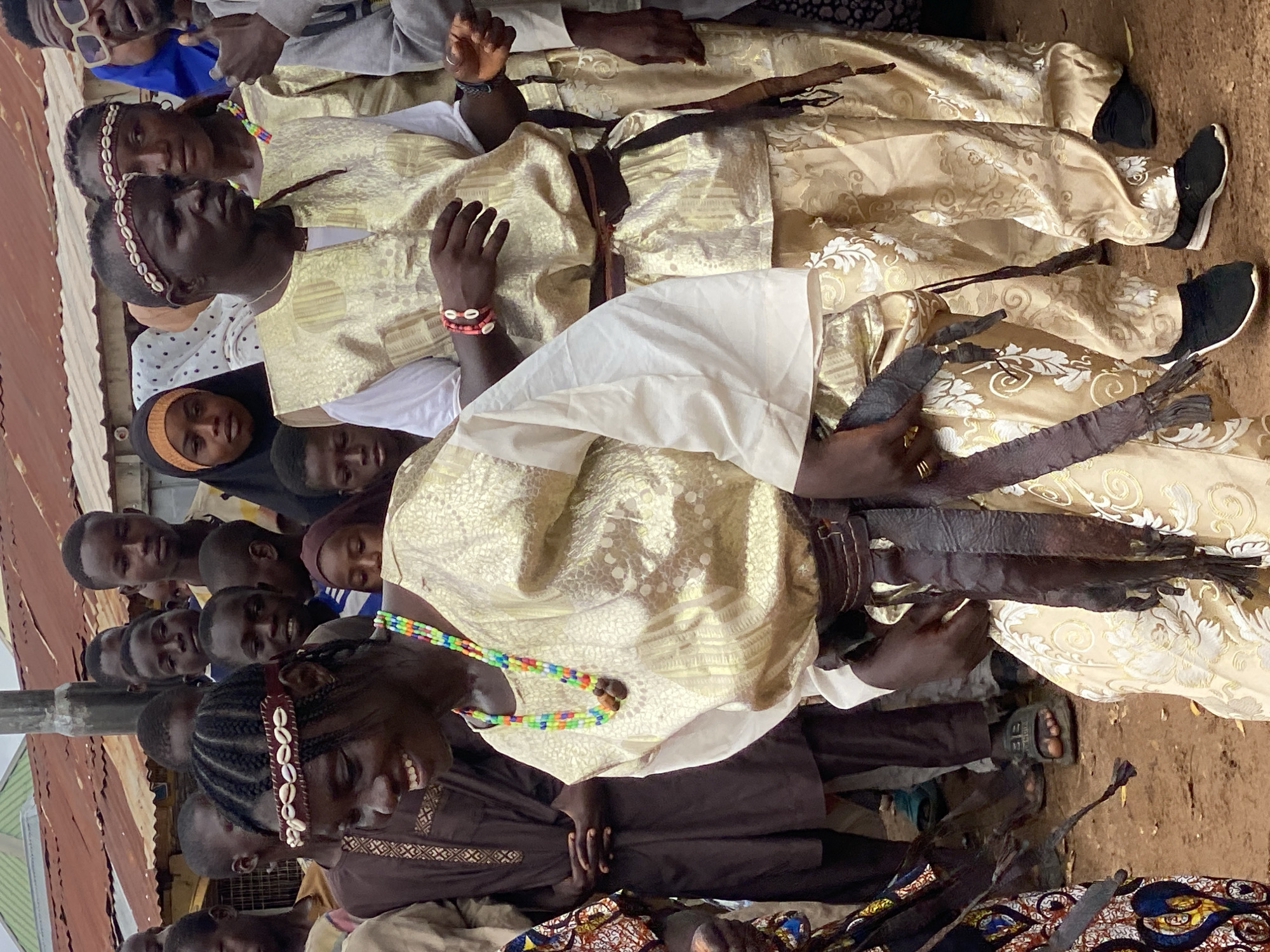 The image captures a group of people, predominantly young men and boys, gathered outdoors in what appears to be a rural setting in Rigachikun, Nigeria. The scene is bustling, with many individuals visible, some in the foreground and others in the background.
Several individuals are dressed in traditional attire. Some wear elaborate, light-colored robes with intricate patterns, possibly for a ceremony or special occasion. One person in the foreground is wearing a dark brown garment with embroidered details and a headband adorned with shells. Another person has a colorful beaded necklace. Some individuals are wearing black shoes, while others are barefoot or wearing sandals.
Objects of interest include what appear to be leather whips or sashes with fringe, held by some of the men. The background shows hints of a rustic environment, with corrugated metal roofing visible, suggesting a simple, possibly impoverished, community. The ground is dusty earth.
The overall mood seems to be one of communal gathering, possibly a celebration or a religious event. The expressions on the faces of the people range from focused to smiling. The lighting suggests it is daytime, with bright sunlight casting shadows. No visible text is present in the image.