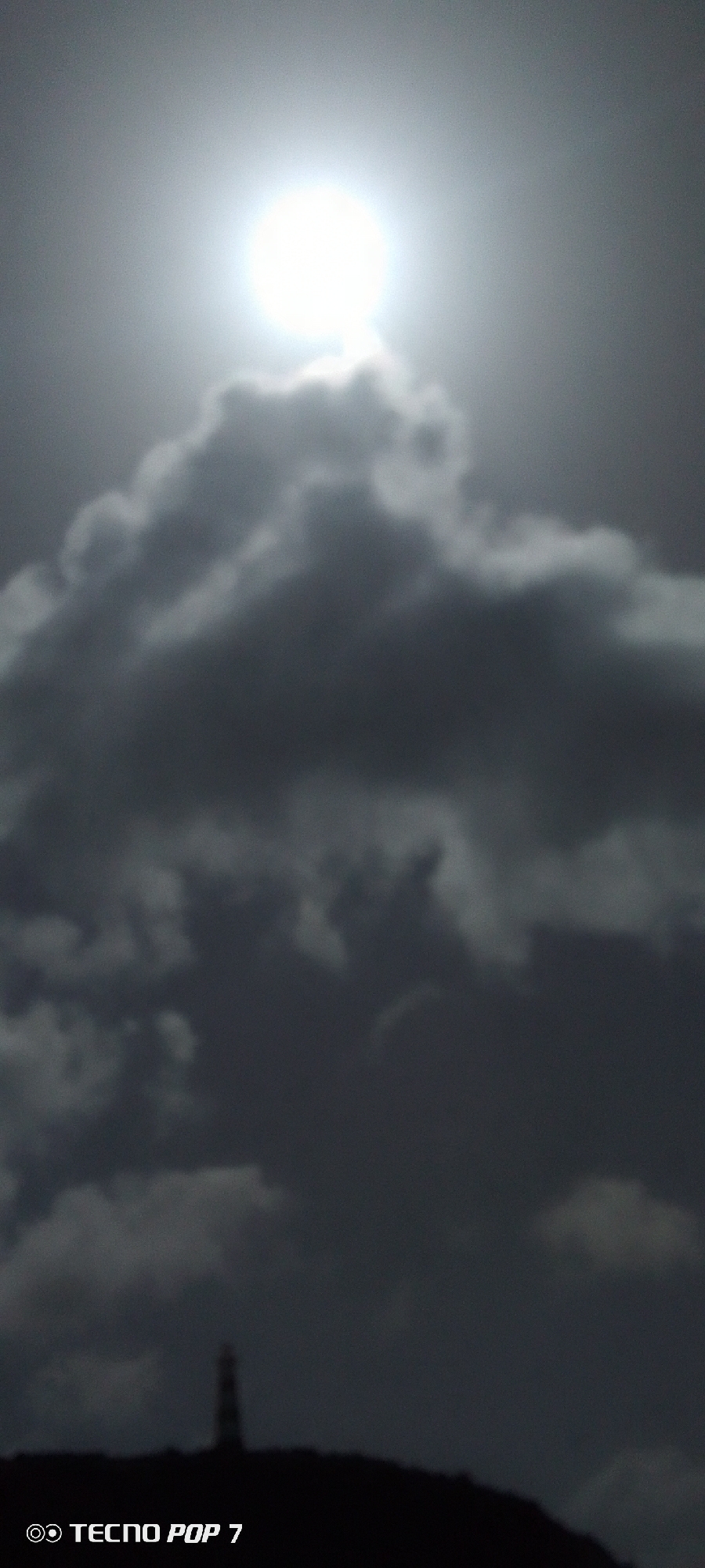 The image depicts a sky filled with dark, heavy clouds obscuring much of the scene. The sun is visible at the top of the frame, its bright light diffused through the clouds. In the lower portion of the image, there is a dark silhouette of land. A tall, striped structure, possibly a lighthouse, is barely discernible on the land. The photo was taken with a TECNO POP 7. This location is in Pedro González, Venezuela.
