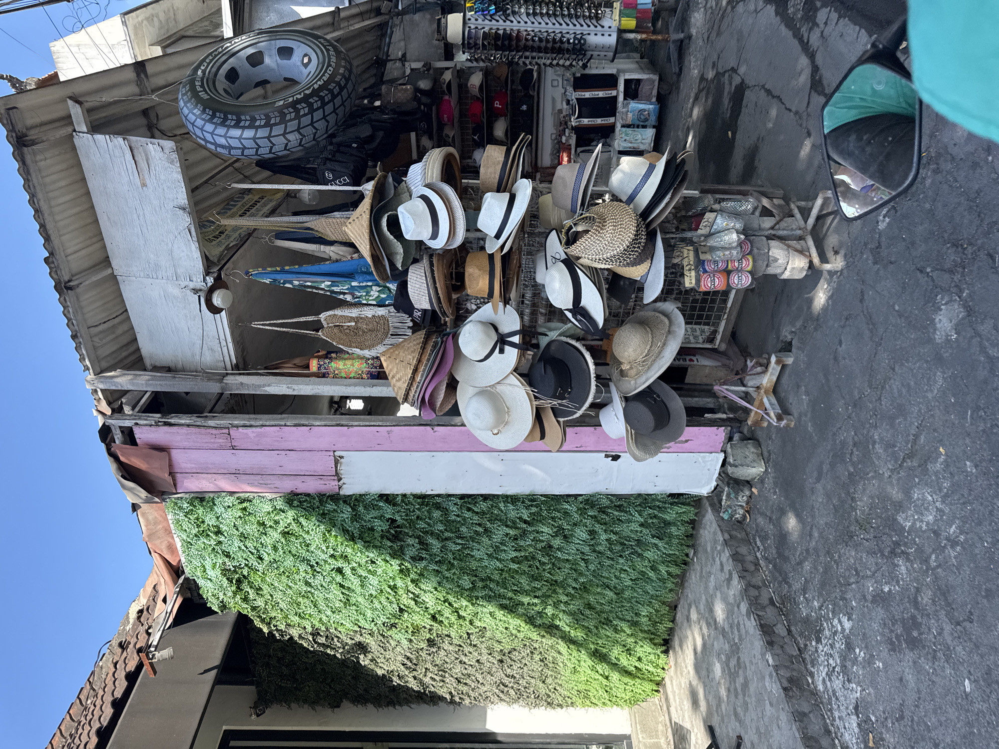 The image depicts a hat vendor in what appears to be a semi-permanent outdoor stall. The stall is constructed from rough-hewn wood, painted pink, white and green. A wide variety of hats, including straw hats and fedoras, are displayed. A tire is hanging above the display. Other items like sunglasses and souvenirs can be seen in the background. The presence of a motorbike mirror suggests the photo was taken from a vehicle. The street is paved. The image was taken in Kuta, Indonesia.
