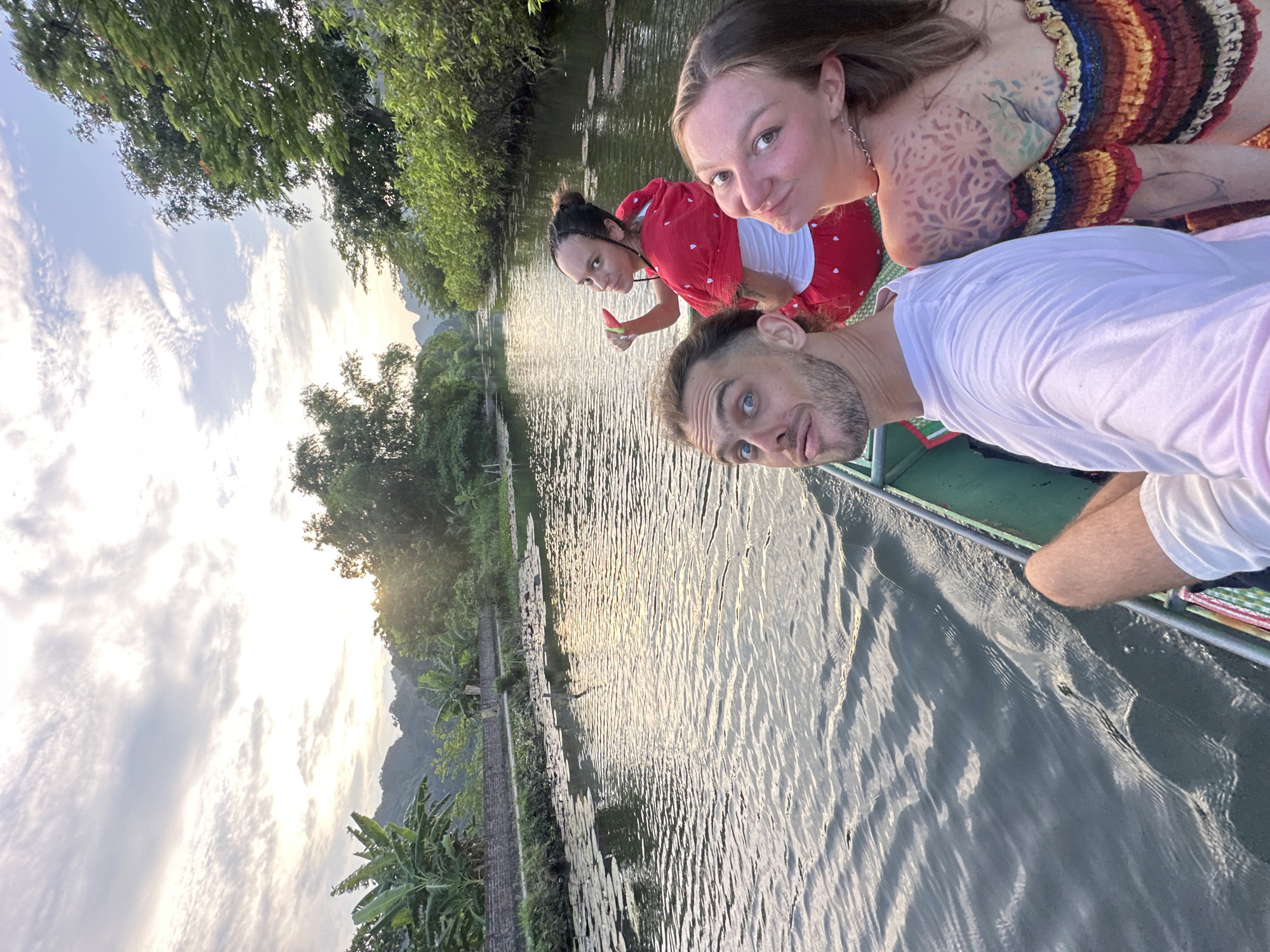 Three people are in a boat on a river. The person in the foreground on the left is a white male with blonde hair and a beard, wearing a white t-shirt. The person in the middle is a white female with dark hair pulled back, wearing a red and white outfit. She is holding a piece of fruit that appears to be watermelon. The person on the right is a white female with blonde hair, wearing a colorful off-the-shoulder top and has tattoos on her shoulder and upper back. The boat is green and moving through the water. The riverbanks are lined with trees and vegetation, and in the distance, there are mountains. The sky is partly cloudy with the sun setting, casting a warm light on the scene. This location is likely in Nam Hoa Lư Ward, Vietnam, known for its scenic karst landscapes and rivers.