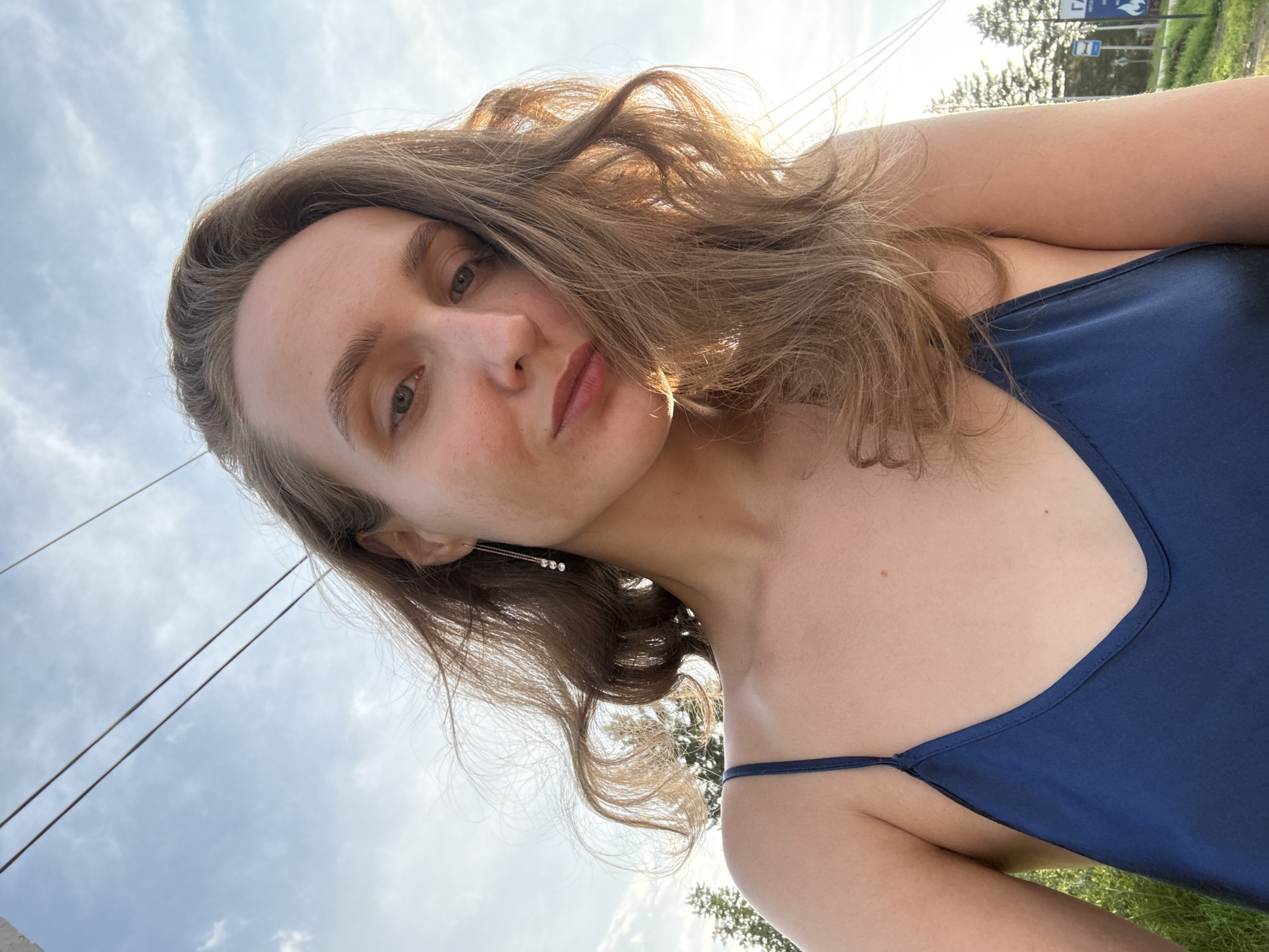 A medium shot selfie shows a young woman with blonde hair and green eyes looking at the camera. She is wearing a dark blue satin camisole. Her hair is windswept, with strands falling across her face. In the background, there is a bright sky with scattered clouds and power lines overhead. To the right of the woman, the edge of a road sign can be seen, with text in Cyrillic script including "ГАЗ" (GAZ) and "Аптека" (Pharmacy). This suggests the location is near a road in a town or village. Considering the request to include the location in Sharkan, Russia, this image likely depicts a scene from that area, possibly taken outdoors near a road with some commercial signage.