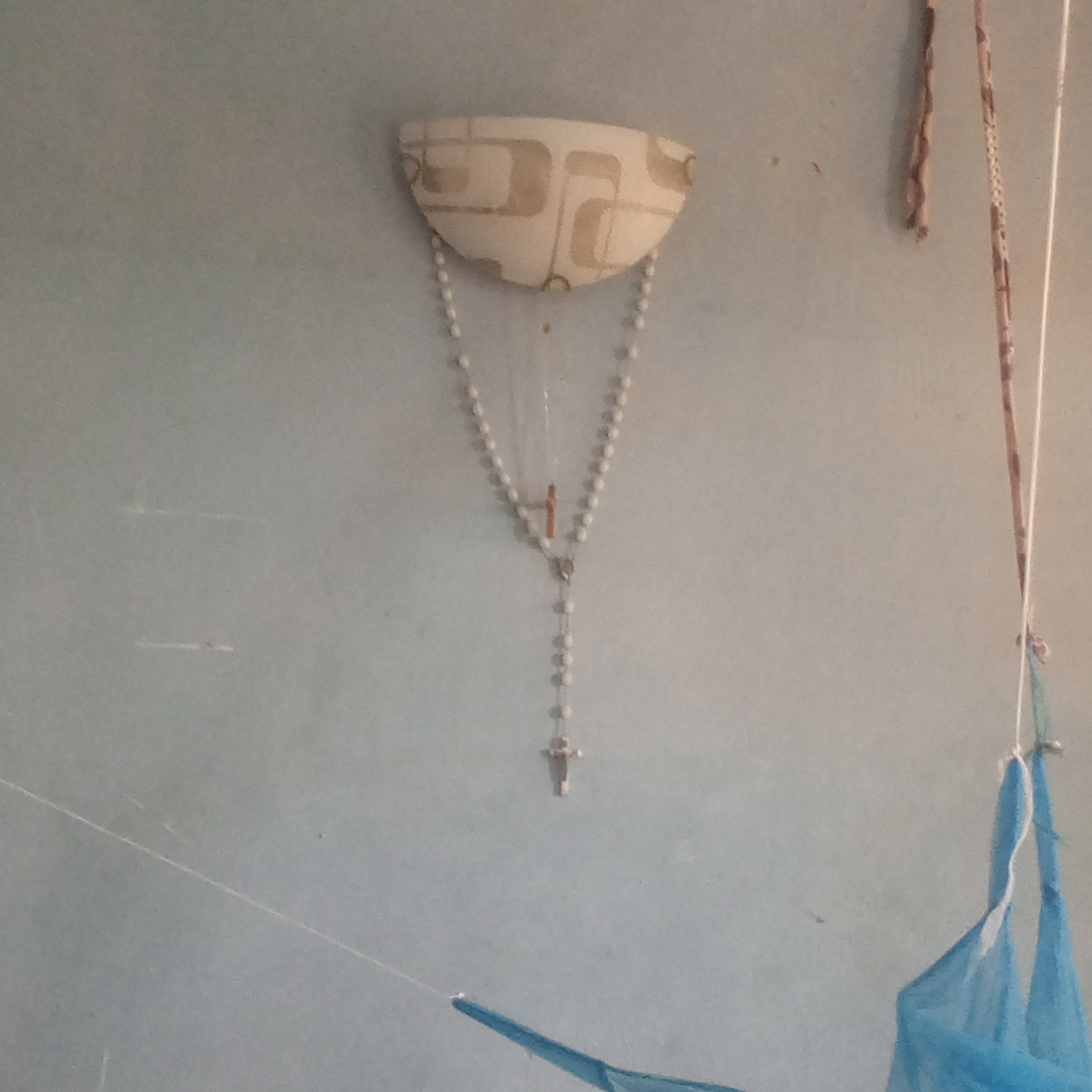 The media file shows a close-up, slightly blurry view of a light blue wall. Prominently featured in the center is a rosary hanging from a decorative sconce. The sconce is semi-circular and has a patterned design of beige rectangles and squares. The rosary itself consists of white beads and a small crucifix. To the right of the rosary, two decorative strings or ribbons hang vertically. One appears to be a twisted brown material, and the other has a repeating pattern of small, square designs. Further to the right, a light blue fabric, possibly part of a mosquito net or curtain, is suspended. The lighting suggests an indoor setting, possibly during the daytime given the general brightness, but no specific time cues are evident. There is no visible text in the image. The overall impression is of a simple, domestic interior.