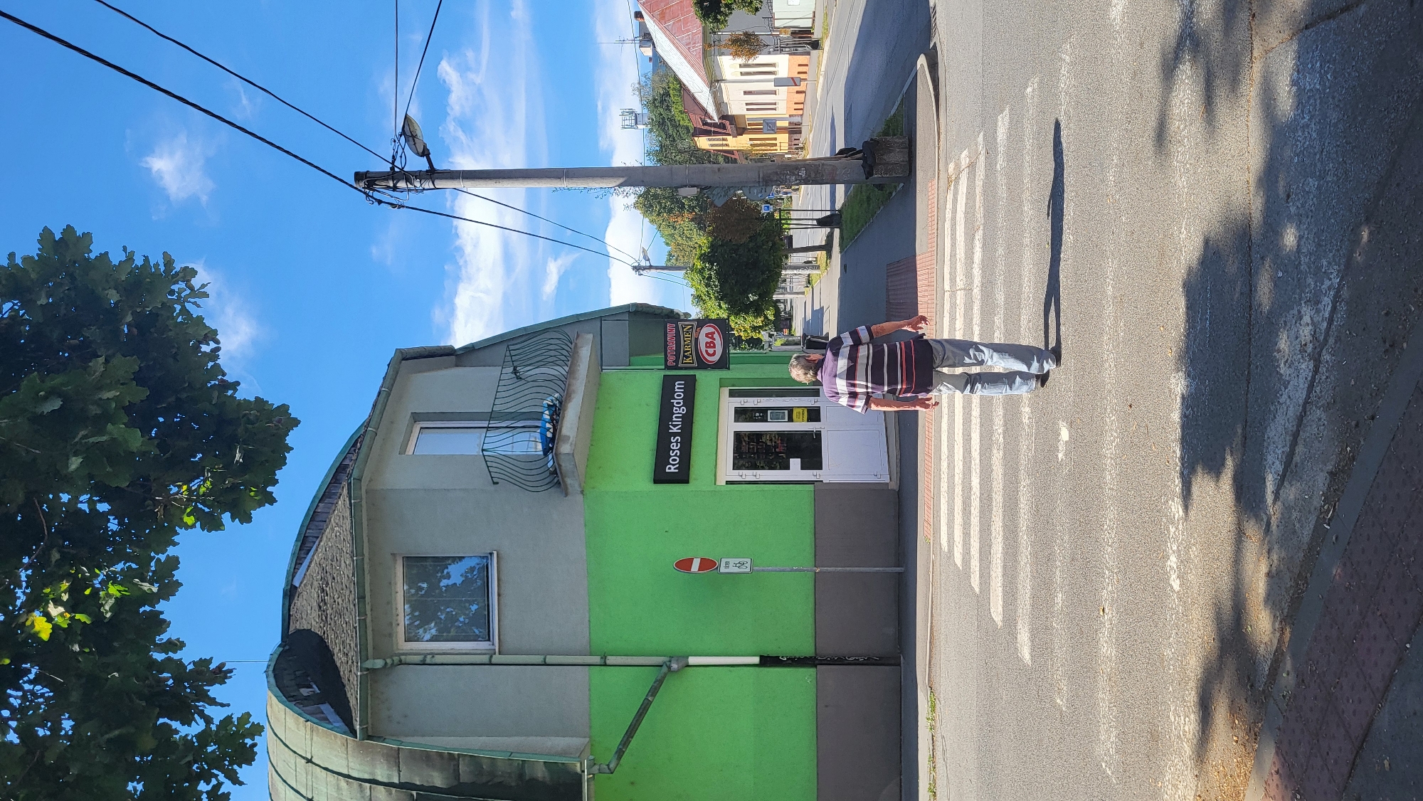 A man walks across a pedestrian crossing on a sunny day in Prešov, Slovakia. The man is wearing a striped shirt and jeans and is walking away from the camera. He is crossing a street in front of a green and gray building with a sign that reads "Roses Kingdom". The building has a distinctive curved, dark green roof and a small balcony with a wrought iron railing. To the right of the building, there are other buildings with colorful facades lining the street, and trees are visible along the sidewalk. A utility pole with power lines and a street lamp stands on the right side of the image. The sky is blue with scattered white clouds.