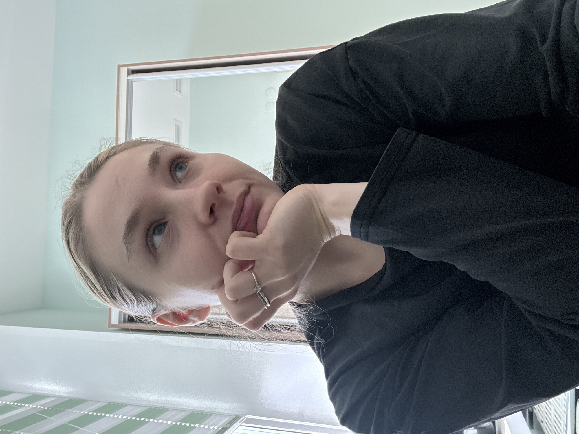The image shows a woman with fair skin, light blonde hair pulled back, and blue eyes. She is wearing a black long-sleeved shirt and a silver ring with a distinctive knot design on her finger. Her head is resting on her hand, and she is looking upwards and slightly to the left. The background consists of a light green wall, a framed mirror revealing another room with white walls, and a partially visible window with a green and white striped blind. The environment suggests an indoor space, possibly a bathroom or bedroom. There is no information to deduce that the location is in the city of Izhevsk, Russia.