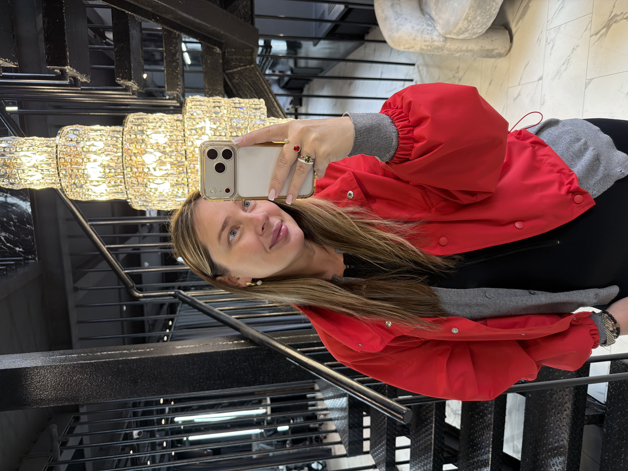 A fair-skinned woman takes a selfie with a smartphone while on stairs in a building in Moscow, Russia. She wears a red jacket over gray and black clothing. On her hand holding the phone are several rings. Behind her, a chandelier hangs above the stairway. A sculpture or furniture element appears blurred in the background. The stairs consist of metal railings and steps. The floor is a light-colored marble.