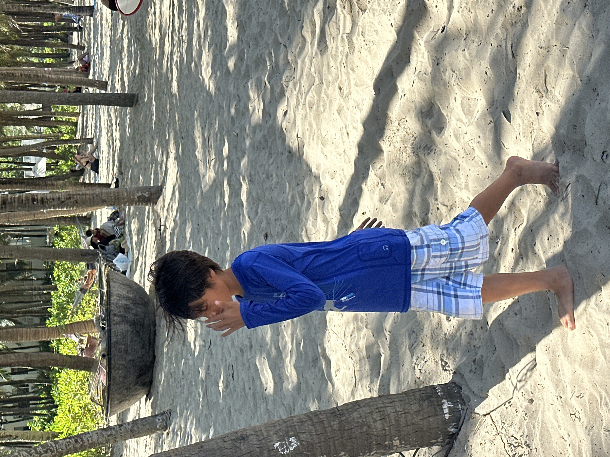 A young boy wearing a blue long-sleeved shirt and plaid shorts stands on a sandy beach in Đà Nẵng, Vietnam, near a palm tree. His right hand is raised to his face, obscuring his nose and mouth. In the background, other people are visible sitting and relaxing under the shade of palm trees, some in hammocks. A large, dark, boat-like object, possibly a coracle or traditional fishing vessel, rests on the sand in the mid-ground. The ground is covered in light-colored sand, with shadows cast by the palm trees overhead.