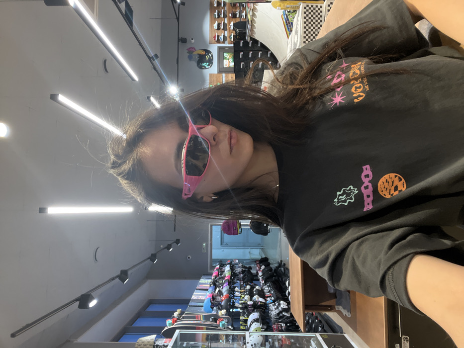 The scene depicts the interior of a retail store specializing in skateboarding and extreme sports apparel and equipment, located in Krasnoyarsk, Russia. The precise address or name of the store cannot be determined from the visual evidence.
A female individual is visible in a mid-shot, appearing to take a self-portrait. She has long, dark, textured hair styled messily and is wearing large, athletic-style sunglasses with bright pink frames and dark, reflective lenses. She is also wearing small, possibly silver or gold-colored hoop earrings and a thin, light-colored necklace. Her attire consists of a dark grey or black short-sleeved t-shirt adorned with multiple colorful graphic designs, including a light green amorphous shape, purple chain links, an orange circular motif resembling a sunset with a checkered pattern, and the brand name "VOLCOM" in orange lettering with pink starbursts and the tagline "WE LIVE FOR WHAT WE LOVE" on the right side of the shirt. Her gaze is directed slightly to her left, away from the immediate camera perspective.
The foreground features a wooden countertop or display table where the individual is positioned. The background reveals the store's interior, organized for merchandise display. To the left, rows of skateboards are vertically displayed against a wall, beneath which are racks holding various helmets (white, black, and patterned) and protective pads. A glass display case is partially visible on the far left. Further back and to the center-left, a doorway with a light blue-painted interior is visible, featuring a partially obscured white "EXIT" sign. A magenta beanie hangs on the wall near this doorway. To the right, additional retail shelving displays shoes, and a prominent wooden skate ramp or quarter pipe with a black and white checkerboard pattern on its top and side is visible. Other skateboards are arranged near the ramp. The ceiling is light grey, equipped with multiple black track lighting fixtures housing tubular fluorescent lights and individual spotlights, providing bright artificial illumination to the space. Large windows are visible on the far left, suggesting natural light penetration from an exterior. The walls are predominantly light grey with some blue accents near the doorway.