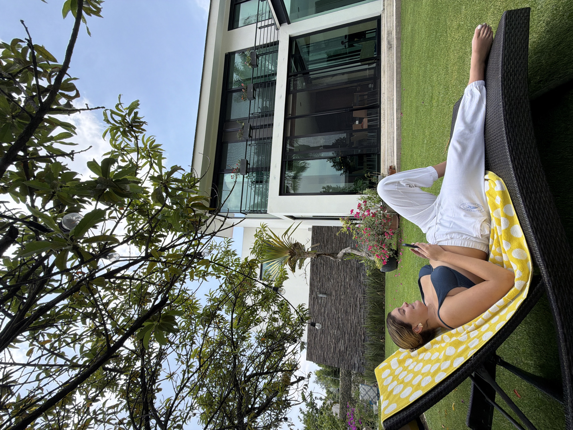 A medium shot of a person reclined on a lounge chair. They are wearing a navy bathing suit top and white pants. They are lying on a yellow towel with white polka dots. They are holding a black smartphone. The person is outside on a patch of green grass. Behind them is a modern two story building with large glass windows. There are trees on the left side of the frame. The sky is blue with light clouds. The city is Unknown, Unknown.