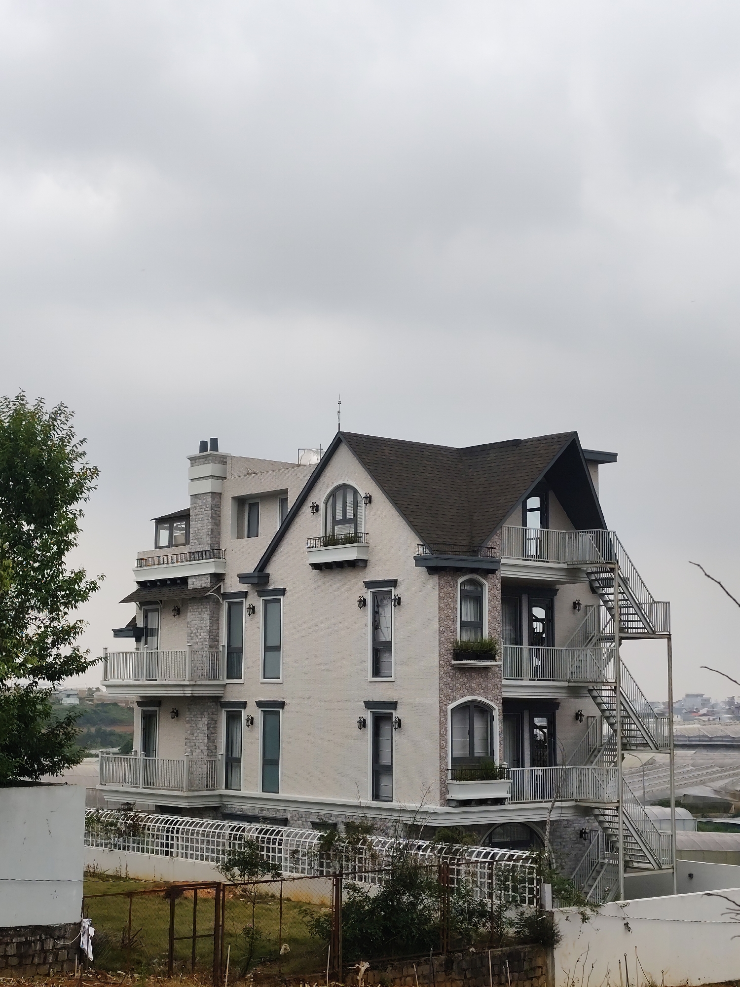 The image shows a multi-story building in what appears to be a residential area. The building has a light-colored exterior with dark-framed windows and several balconies with metal railings. A prominent metal staircase is attached to one side of the building. There is a chimney on the left side and a dark-colored roof. The sky is overcast. Fences and some vegetation are visible in the foreground. In the distance, there appears to be a city landscape, possibly consisting of greenhouses, consistent with the location of Lam Vien - Da Lat, Vietnam. There are no people visible in the image.