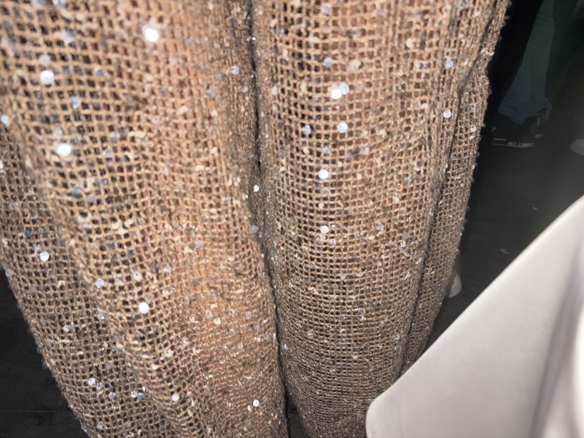 A close-up, low-angle, slightly blurry shot shows a shimmering, loosely woven, brown fabric with scattered sequins. The fabric appears to be draped, creating folds and shadows. To the right of the fabric, a section of a light-colored, curved object is visible, possibly a piece of furniture or clothing. In the background, a dark area with hints of clothing and footwear suggests an indoor setting. The image does not provide any specific visual cues to place it within the city of Dénia, Spain.