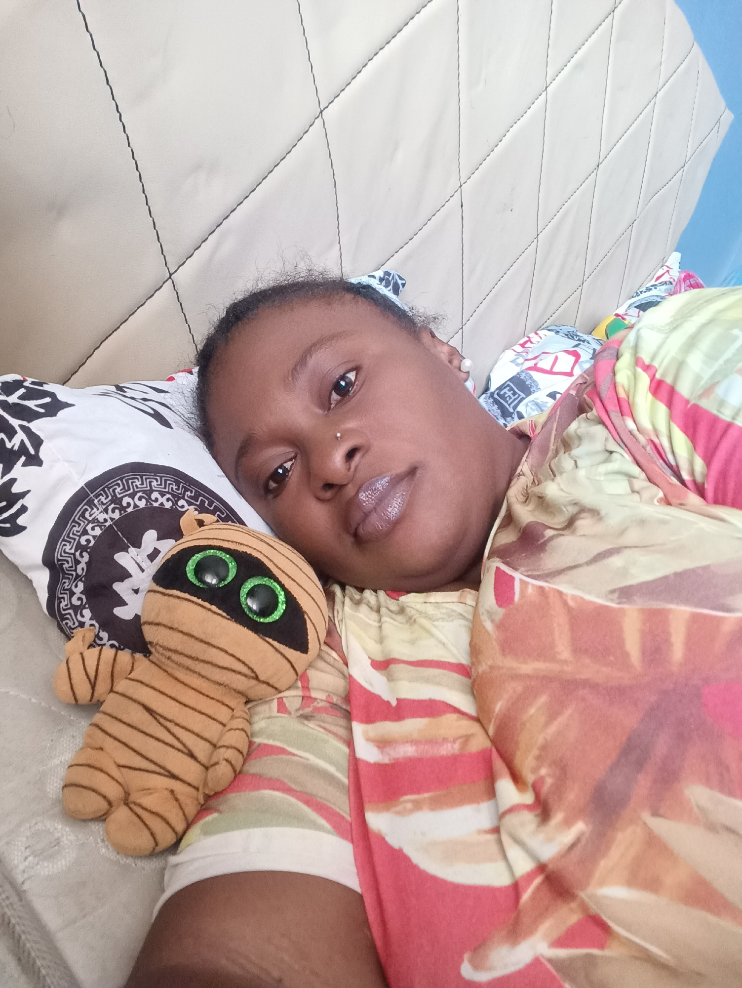 A person with dark skin and short dark hair is lying on a bed. They have a nose piercing. A stuffed mummy toy with large green eyes is next to them on a pillow with a black and white design. The bed has a beige quilted headboard. The person is covered with a multicolored blanket. The location is Jos, Nigeria.