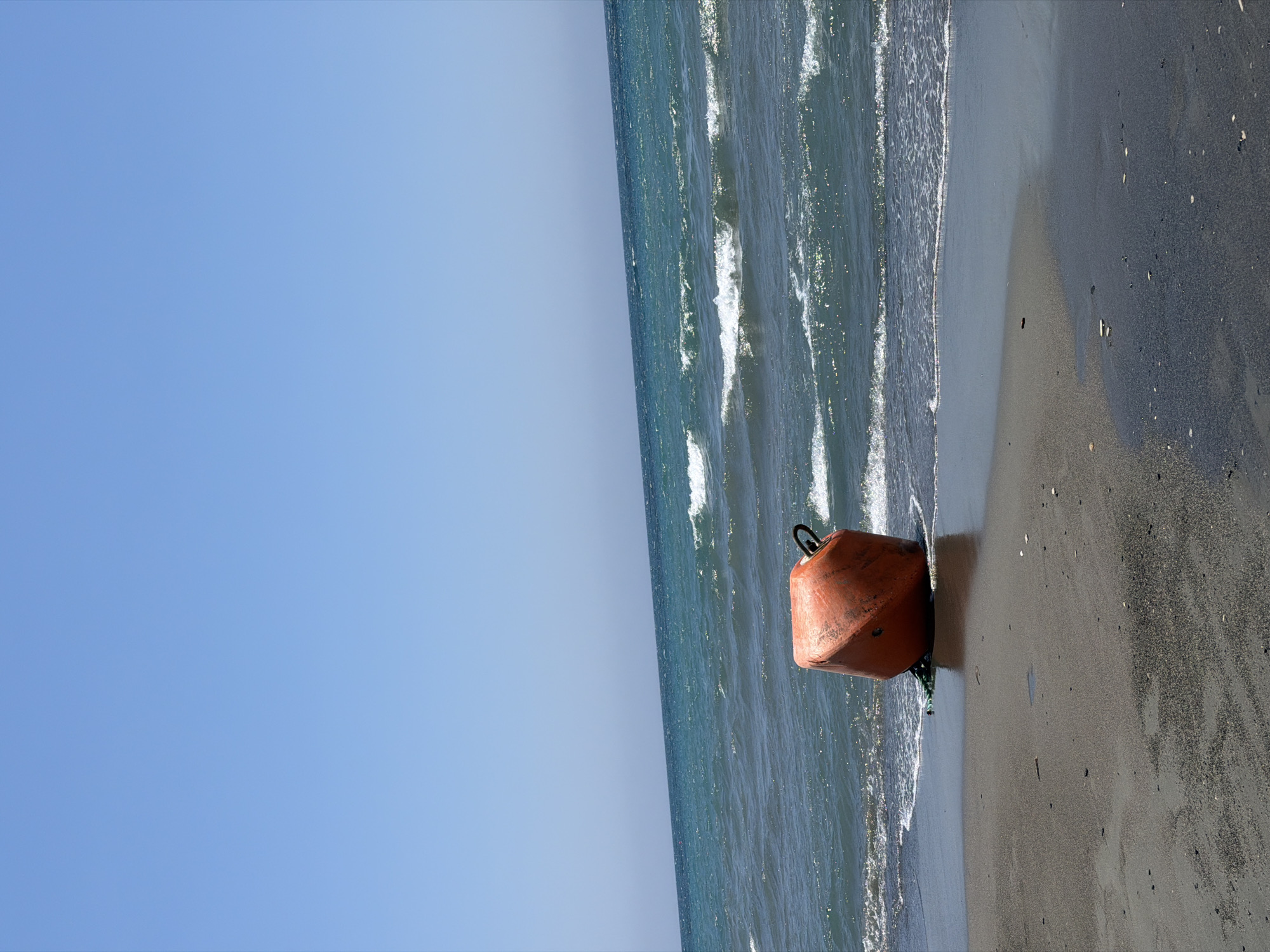A bright orange, cubical buoy with a metal loop on top rests on a dark sandy beach. The buoy is partially submerged by shallow, foamy waves of a dark blue sea under a clear, pale blue sky. The horizon line is visible in the distance, separating the sea from the sky. The beach is wet in the foreground, reflecting the sky, and drier sand with scattered small pebbles further back. The scene is devoid of people. The location is the coast of Meneou, Cyprus.