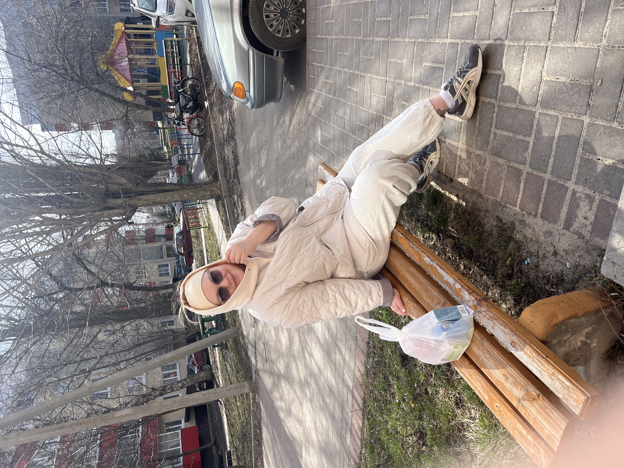 A person wearing a light-colored quilted jacket, sweatpants, sneakers, a head covering, and sunglasses is lying on a wooden bench. Next to the person is a clear plastic bag containing various items. The bench is situated on a paved walkway, adjacent to grass and a parking area where a silver car is partially visible. In the background are leafless trees, multi-story buildings, and a playground. The location is Usman, Russia.