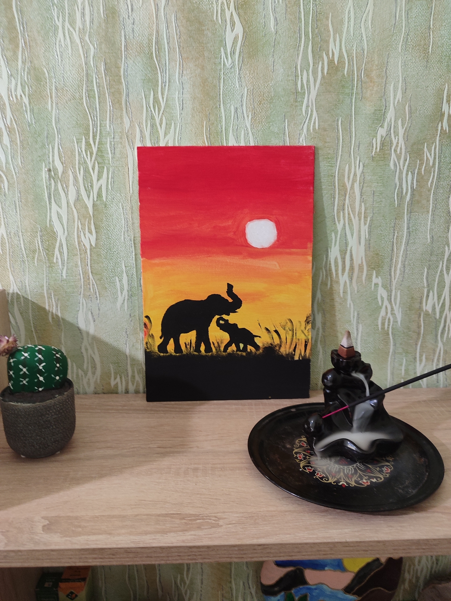 A still life image captures a painting of elephants silhouetted against a sunset, an incense burner emitting smoke, and a decorative cactus. The painting, featuring a red and orange gradient sky with a white sun, depicts a large elephant with a calf standing amidst stylized grass. To the left of the painting is a small, green, decorative cactus in a textured gray pot. To the right of the painting is a black ceramic incense burner shaped like a figure, with a cone incense stick smoldering, producing a stream of white smoke that curls over the burner and onto a decorative black plate with red and yellow floral accents. The objects are placed on a light-colored wooden shelf. The background is a textured green wallpaper with a repeating pattern of thin, white, vertical lines resembling branches or reeds. This scene is located within a room in the city of Lozuvatka, Ukraine.