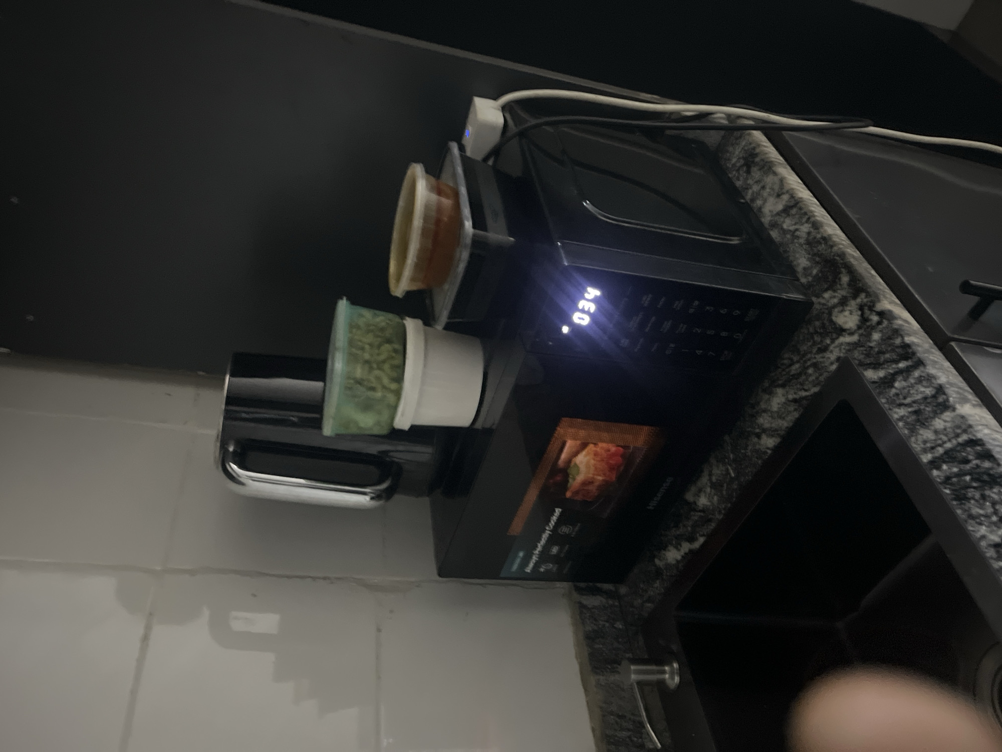 A black Hisense microwave oven sits on a granite countertop next to a black sink. The microwave displays "034" and an image of food. On top of the microwave are two containers of food, one green and one orange. A kettle is to the left of the food containers. The background is a white tiled wall with a shadow on it. The scene is in a kitchen in Karum, Nigeria.