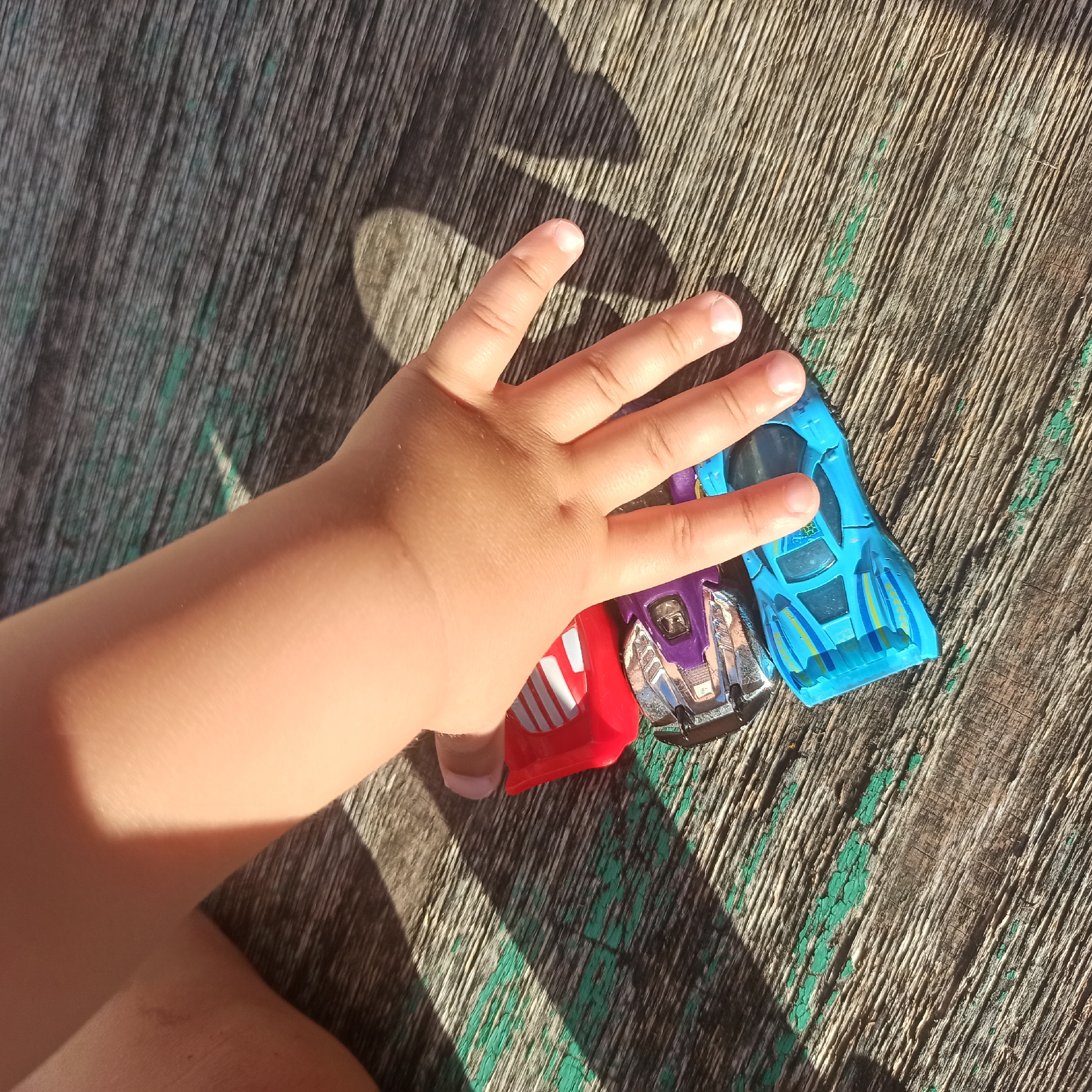 The image shows a close-up view of a child's hand resting on a stack of toy cars. The cars are positioned on a weathered wooden surface with visible green paint chipped away in places, suggesting an outdoor environment. Sunlight casts shadows across the scene, highlighting the texture of the wood and the contours of the child's hand. The cars appear to be a red one on top, followed by a purple and silver car, and finally a blue car at the bottom. The child's fingers are gently covering parts of the purple and blue cars. No specific location in Nizhny Novgorod, Russia, can be determined from the provided image.