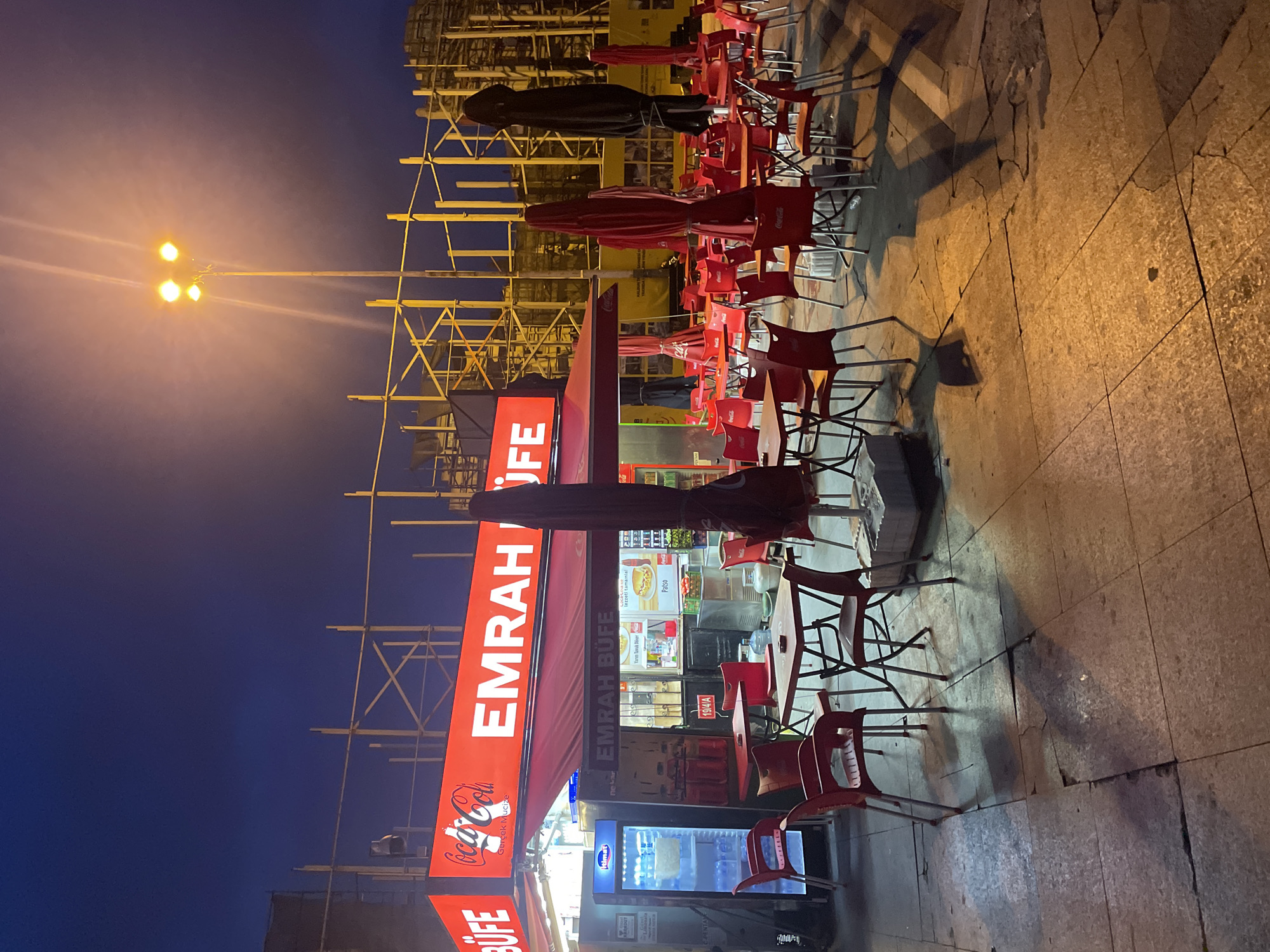 The image shows an outdoor seating area of a food establishment, likely a "büfe" (kiosk or small eatery), named "EMRAH BUFE" as indicated by a prominent red illuminated sign. The establishment is situated on a paved sidewalk with visible cracks and unevenness. In the background, there appears to be ongoing construction or renovation work, indicated by scaffolding and building materials.
The seating area consists of numerous red plastic chairs and tables, many of which are empty. Several large, closed umbrellas, some red and one black, are deployed over the tables, suggesting an attempt to shield the area from weather or perhaps for nighttime ambiance. A refrigerated display case, labeled "Himel," is visible to the left of the entrance, containing drinks. Another smaller display case inside the establishment shows food items. Bottles of Coca-Cola are visible both in the display case and on shelves behind the counter.
A street lamp with multiple bulbs illuminates the scene from the upper right, casting long shadows across the pavement. The sky is a deep twilight blue, indicating it is either early morning or late evening. The overall impression is that of a street-level business in a Turkish city, possibly Kadıköy, given the establishment's name and context. There are no people visible in the immediate seating area or around the establishment. The environment appears to be a public urban space.